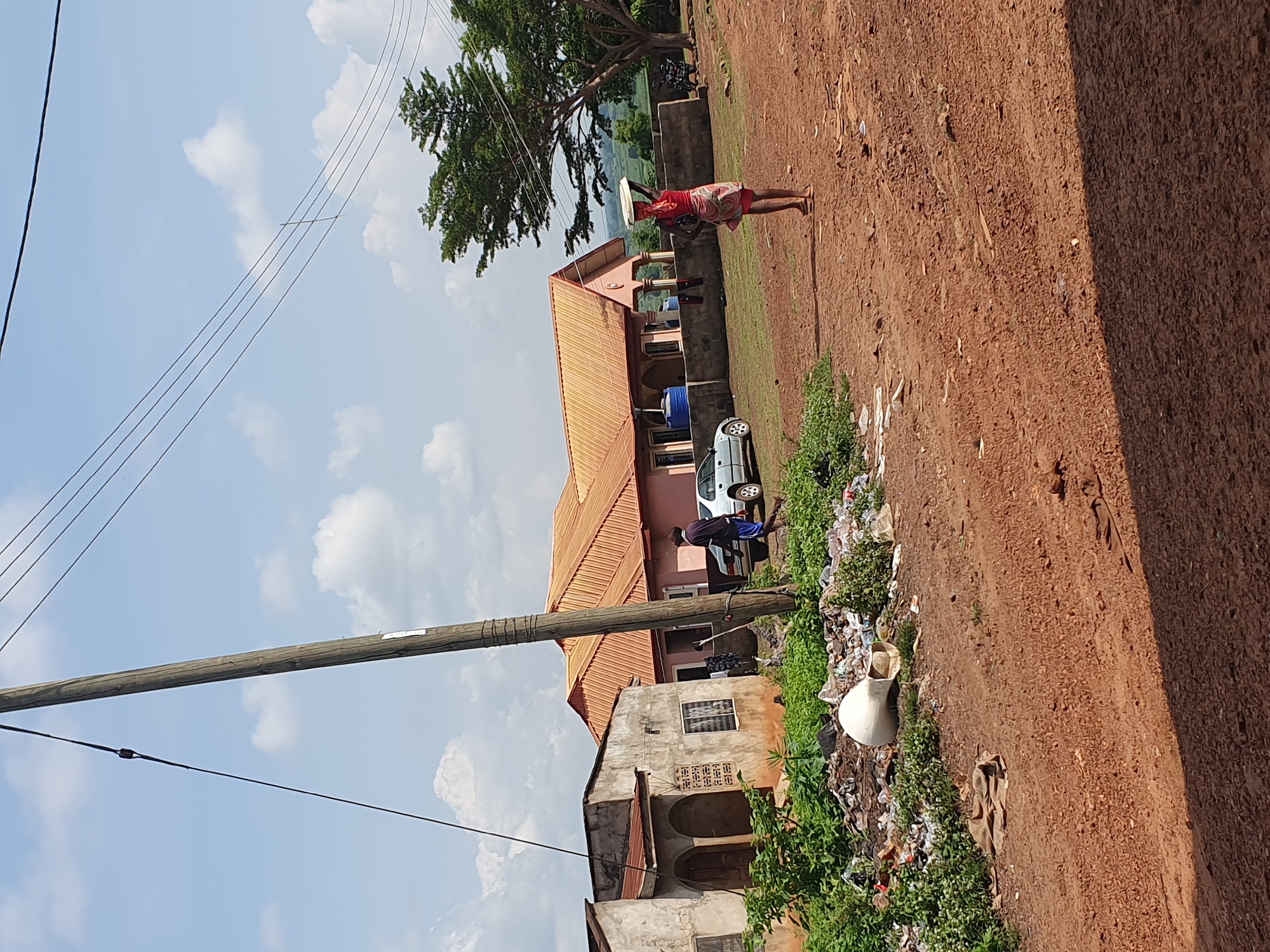 The image captures a vibrant, sunlit street scene in what appears to be a rural or semi-urban area of Idah, Nigeria. The dominant feature is a wide, unpaved dirt road, characterized by its reddish-brown earth, which stretches diagonally across the frame from the foreground towards the background.
In the middle ground, a woman walks barefoot along the dirt path, carrying a large, flat, light-colored object expertly balanced on her head. She is dressed in a red top and a colorful patterned skirt. Behind her, a large tree with lush green foliage provides shade, and a substantial building with a reddish-orange corrugated roof and light-colored walls is visible. A silver car is parked near this building, with a person standing close by.
To the left of the road, a prominent utility pole rises into the bright blue sky, carrying multiple power lines that crisscross the upper left quadrant of the image. Further down, closer to the foreground, is another, older-looking building with a grey, weathered facade. Along the edge of the dirt road, to the left, there's a strip of green vegetation intertwined with a considerable amount of scattered trash and debris, including plastic bottles and other discarded items. An overturned white pot lies amidst this refuse.
The sky is a clear blue with scattered white, cumulus clouds. The strong sunlight indicates it is daytime, likely mid-morning or afternoon, casting visible shadows on the ground. In the distance, beyond the buildings and trees, hints of green hills or a natural landscape can be seen. No clear, readable text is visible on any objects within the image.