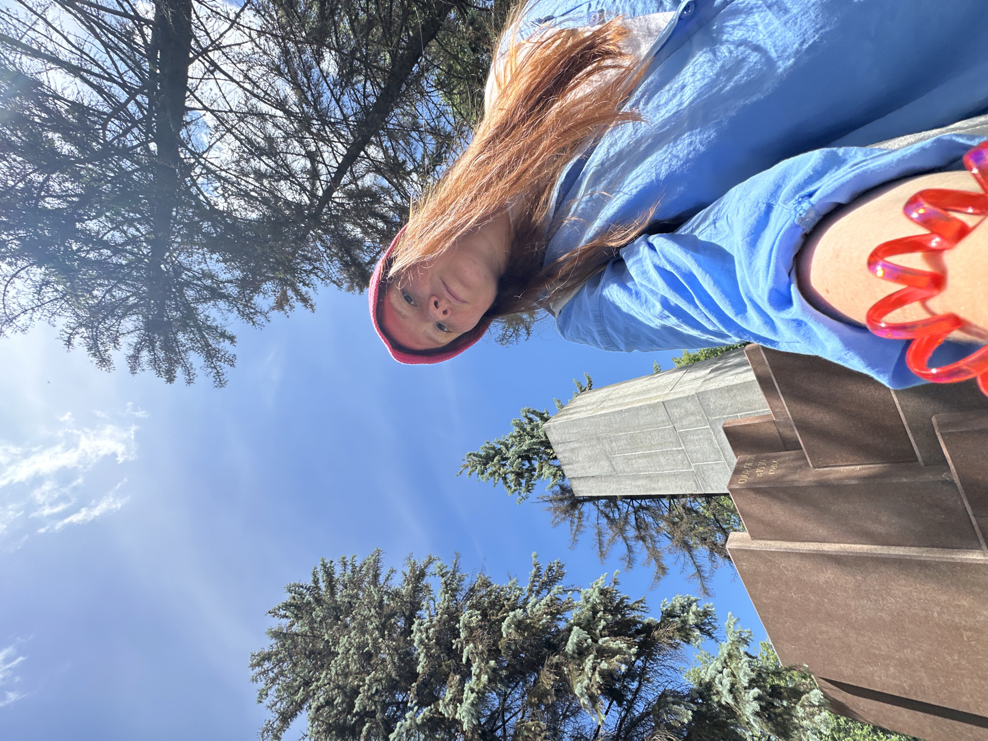 The image is a bright, daytime shot taken from a low, upward-looking angle, featuring a person in the foreground against a backdrop of sky, trees, and architectural structures.
The main subject is a woman with long, reddish-brown hair, wearing a light blue long-sleeved shirt and a red beanie. She is looking directly downwards at the camera with a neutral expression. A bright, iridescent red, spiral-shaped bracelet is visible on her right forearm.
The setting appears to be an outdoor park or memorial area. The sky is a clear, vibrant blue with scattered white clouds, indicating sunny weather. Various evergreen trees with dense, bluish-green needles are visible, along with some trees featuring sparser, darker branches.
In the mid-ground, two distinct architectural structures are prominent. One is a tall, grey, block-like structure with a tiled or textured surface. Adjacent to it is a multi-layered, stepped structure made of reddish-brown material, possibly weathered metal or stone. Visible Cyrillic text on the brown structure reads "СКОРБЬ" (Sorrow/Grief), "ПАМЯТЬ" (Memory), and the years "1941" and "1945". This text strongly suggests the structures are part of a war memorial, specifically commemorating the Great Patriotic War (World War II).
The scene is captured in Nizhny Novgorod, Russia, at what appears to be a solemn and well-maintained memorial site. The unusual perspective adds a dynamic element to the capture of the person and their surroundings.