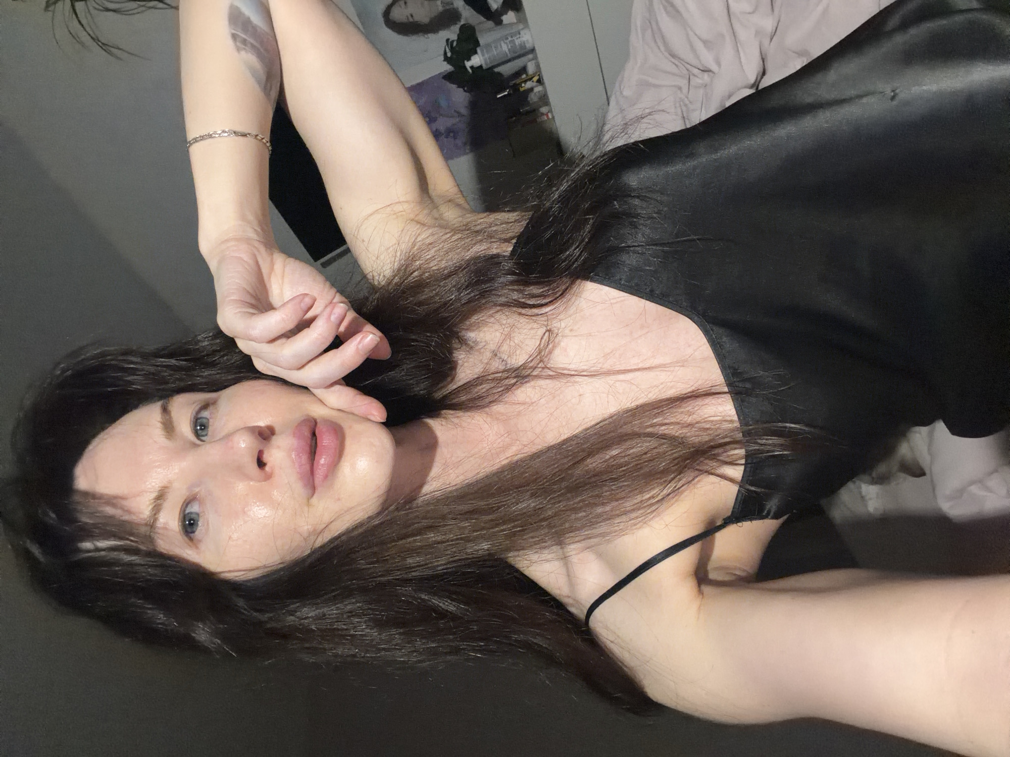 A woman lies down wearing a black satin top. She is pale, with blue eyes and brown hair, and has a bracelet on her left wrist and a tattoo on her left forearm. The background includes bedding and a poster. The location is not specified.