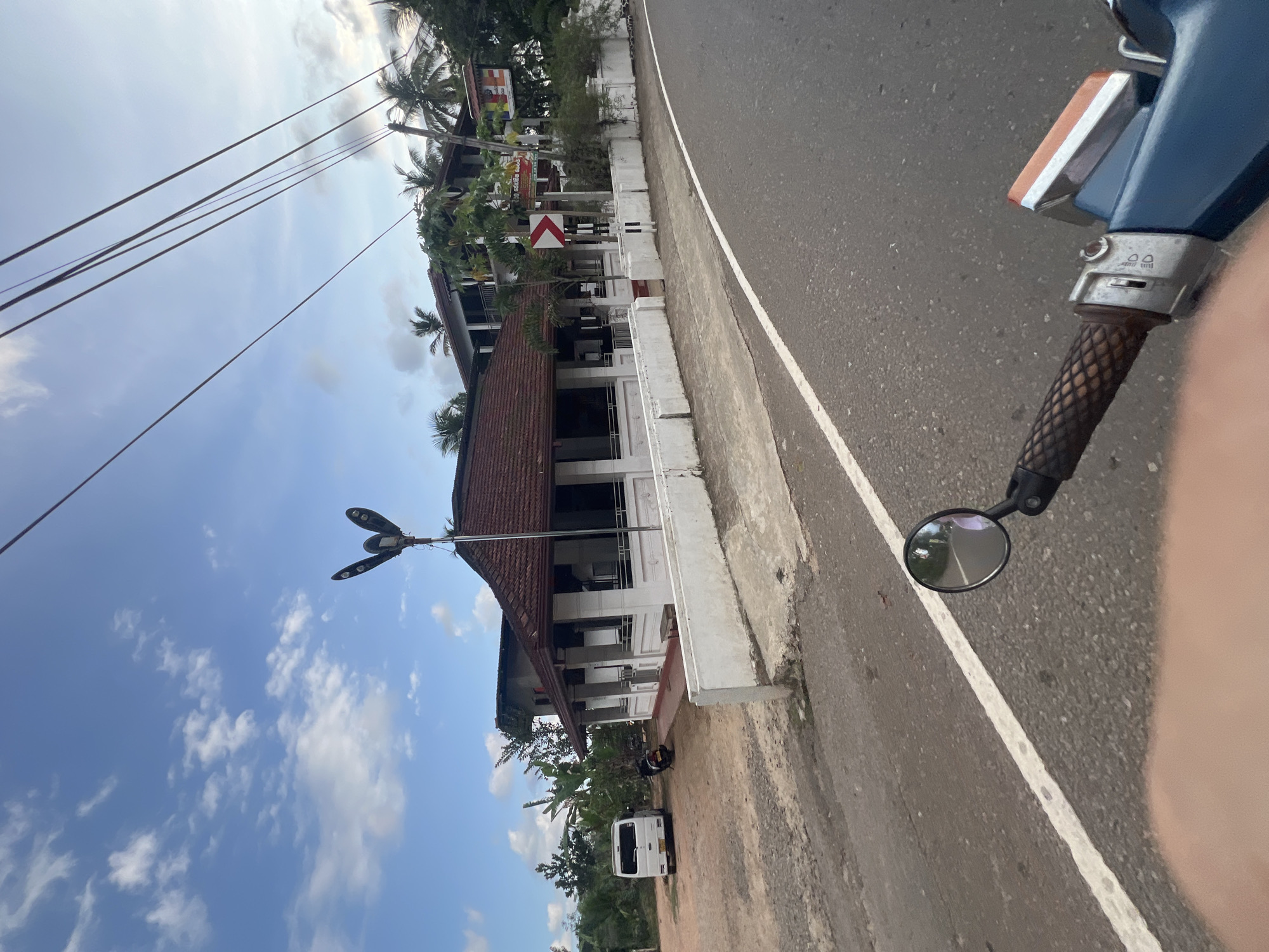 The image captures a roadside scene near Angulugaha Junction, Sri Lanka. The view is taken from the perspective of someone on a moving vehicle, likely a scooter, as part of the handlebar and a side mirror are visible on the right side of the frame.
To the left, a two-story building with a red tile roof stands parallel to the road. A white wall runs along the edge of the building's property, bordered by a narrow dirt area and the road. A white van is parked in the dirt area near the building, slightly further down the road. A three-armed street light fixture is also located by the building.
The road itself is asphalt with a white dividing line. Palm trees and other greenery can be seen in the background, suggesting a tropical environment. Power lines stretch across the top left portion of the image against a partly cloudy sky. The overall scene appears to be a typical roadside setting in a semi-urban or rural area of Sri Lanka.