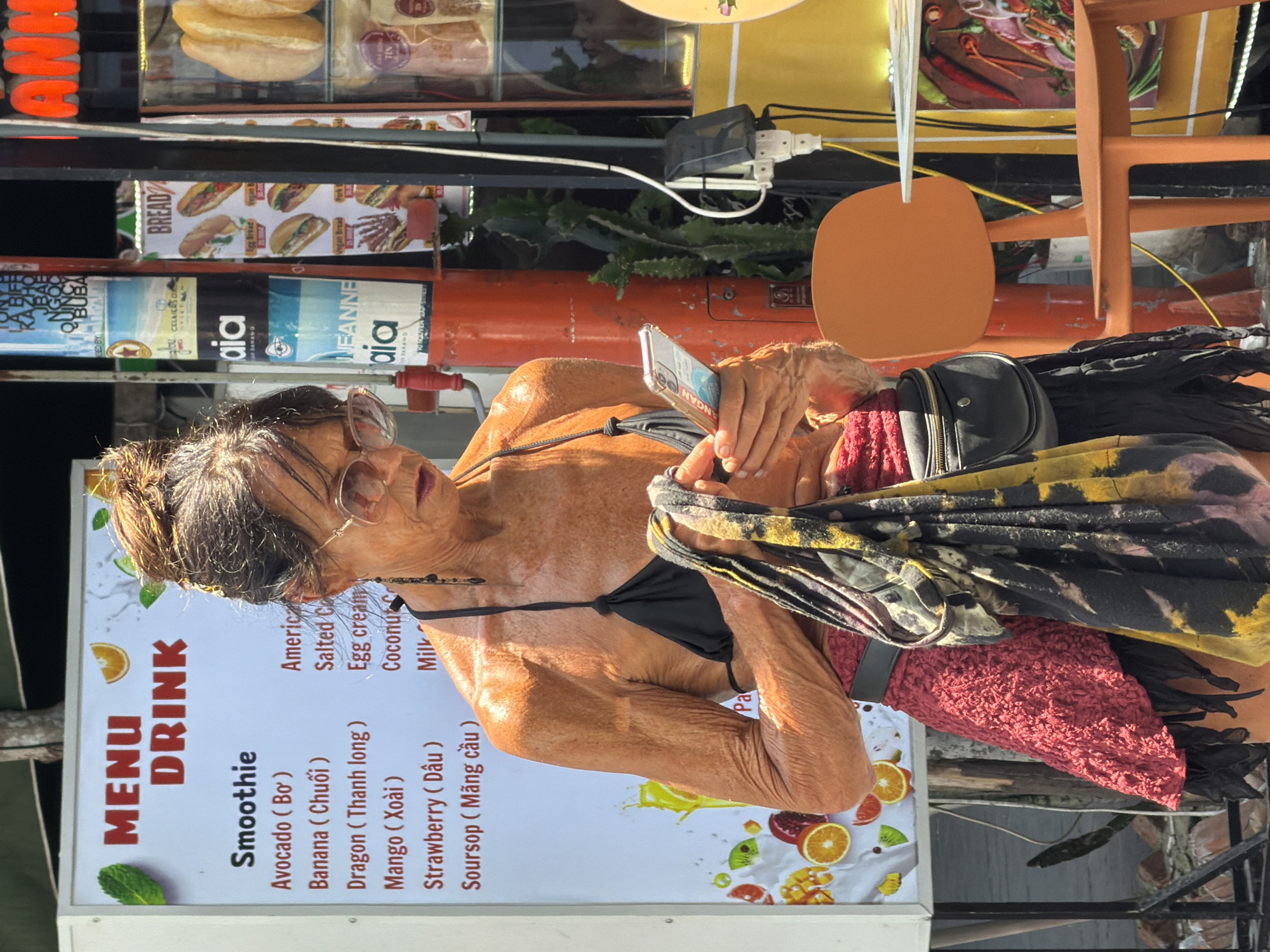 In the city of Đà Nẵng, Vietnam, an elderly woman with sunglasses wears a black bikini top and holds a smartphone. She stands next to a food cart that features a menu of drinks, including smoothies and other options such as "Salted Coffee" and "Egg cream." The cart also displays an advertisement for "BREAD" with images of various sandwiches. Behind the cart are additional items, including a tank, a chair, and an overhead display case containing packaged foods.