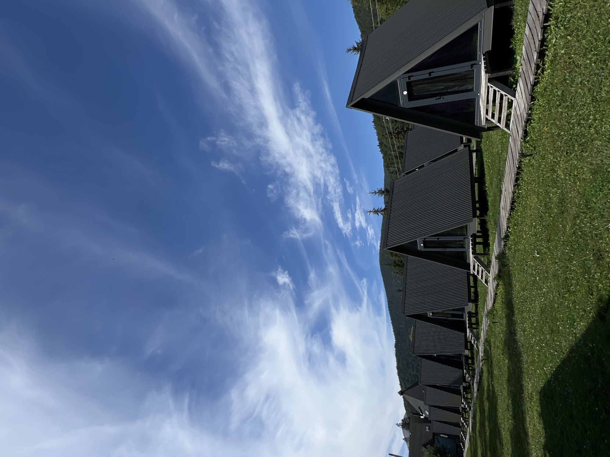 In Vorokhta, Ukraine, a row of modern, dark gray A-frame cabins stands on a grassy lawn under a partly cloudy blue sky. A wooden boardwalk path runs along the front of the cabins, with small wooden stairs leading up to each entrance. The cabins are identical, with corrugated metal roofs and dark-framed glass doors. In the background, a dense, dark green forest covers a sloping hillside, with a few scattered pine trees also visible on the lawn. The overall scene suggests a peaceful, natural setting.