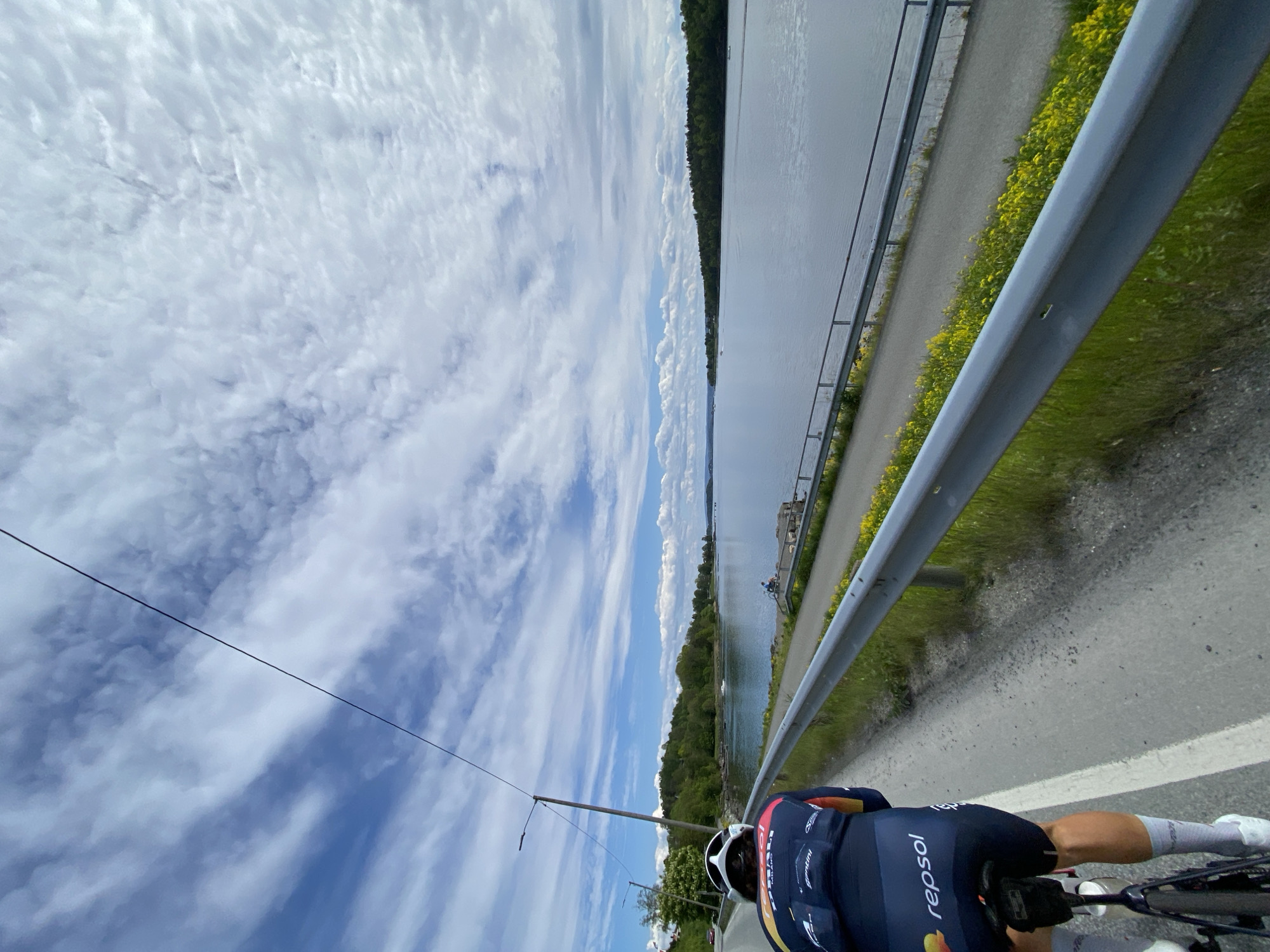 A cyclist is riding on a paved path adjacent to a body of water, possibly the Oslofjord, in Asker, Norway. A metal guardrail separates the path from a grassy area with yellow flowers along the water's edge. The cyclist is wearing a dark blue cycling jersey with "repsol" printed on the back, along with a white helmet and white socks. The sky is partly cloudy. Overhead electrical wires are visible. A secondary path or embankment is visible on the opposite side of the water. Distant trees and structures are present on the other side of the Oslofjord.