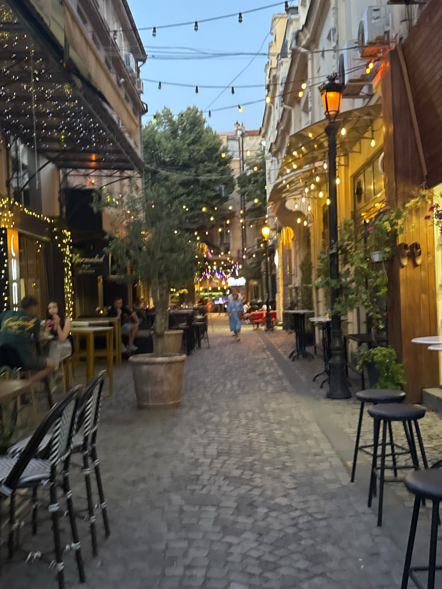 The image depicts a narrow cobblestone street in Plovdiv, Bulgaria, lined with buildings adorned with outdoor seating areas and string lights. The scene is set in the early evening, as suggested by the soft lighting from street lamps and numerous string lights illuminating the facades of the buildings. On the left side, a restaurant or cafe features tables and chairs, with patrons seated and enjoying their time. An olive tree in a large pot stands prominently in the foreground. On the right side, more outdoor seating is visible, including high stools around a table, and a decorative lamppost. A woman in a light blue dress is walking down the street, appearing to be the only person actively moving through the scene. The overall atmosphere is one of a charming and inviting European street.