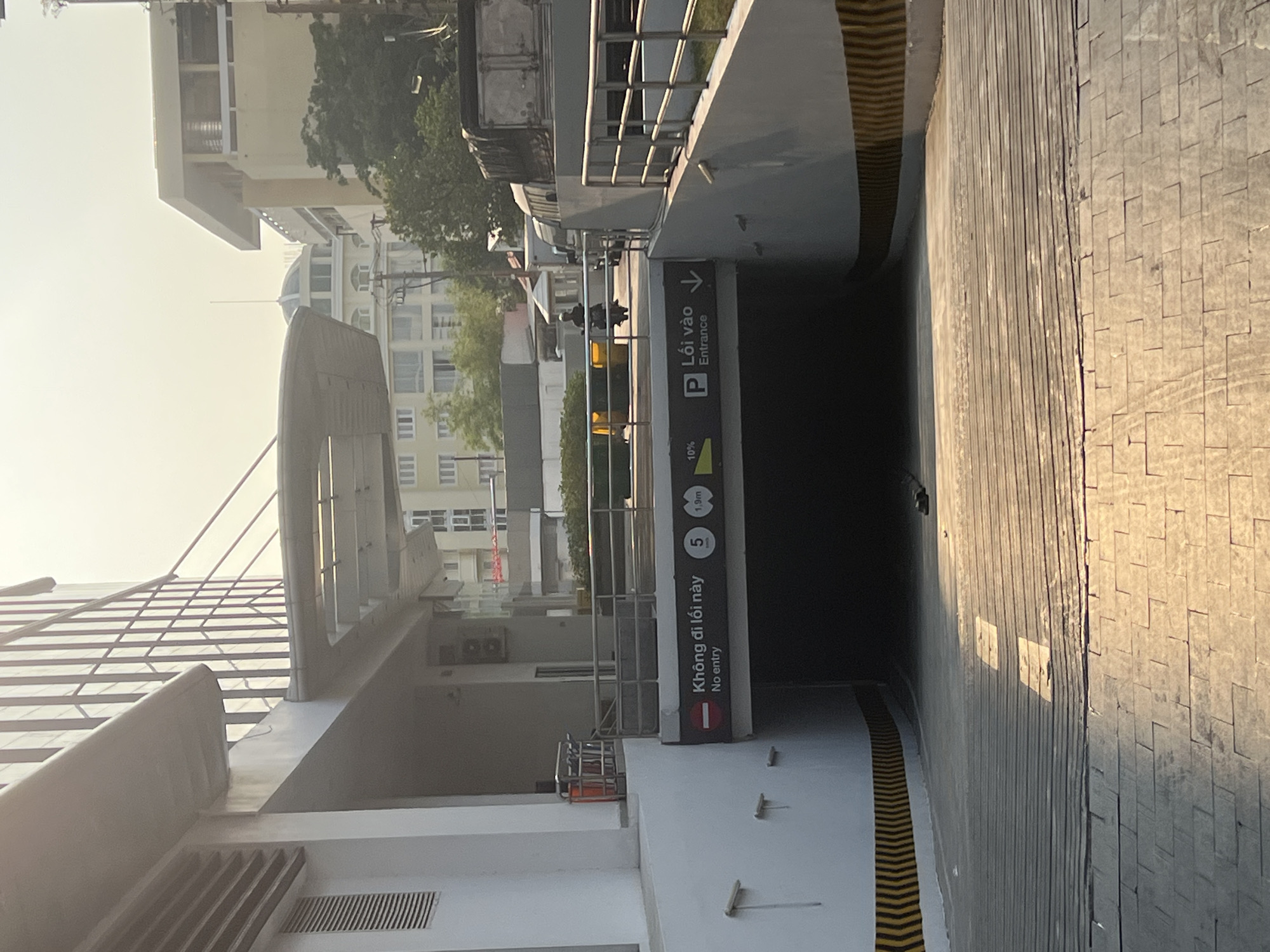 The image shows the entrance to an underground parking garage in Nha Trang, Vietnam. A sign indicates "Lối vào Entrance" with a parking symbol and an arrow, along with a height restriction of 1.9 meters. Another sign indicates "Không đi lối này No entry" with a red bar. The garage entrance is on a paved inclined road. In the background, buildings and trees are visible.