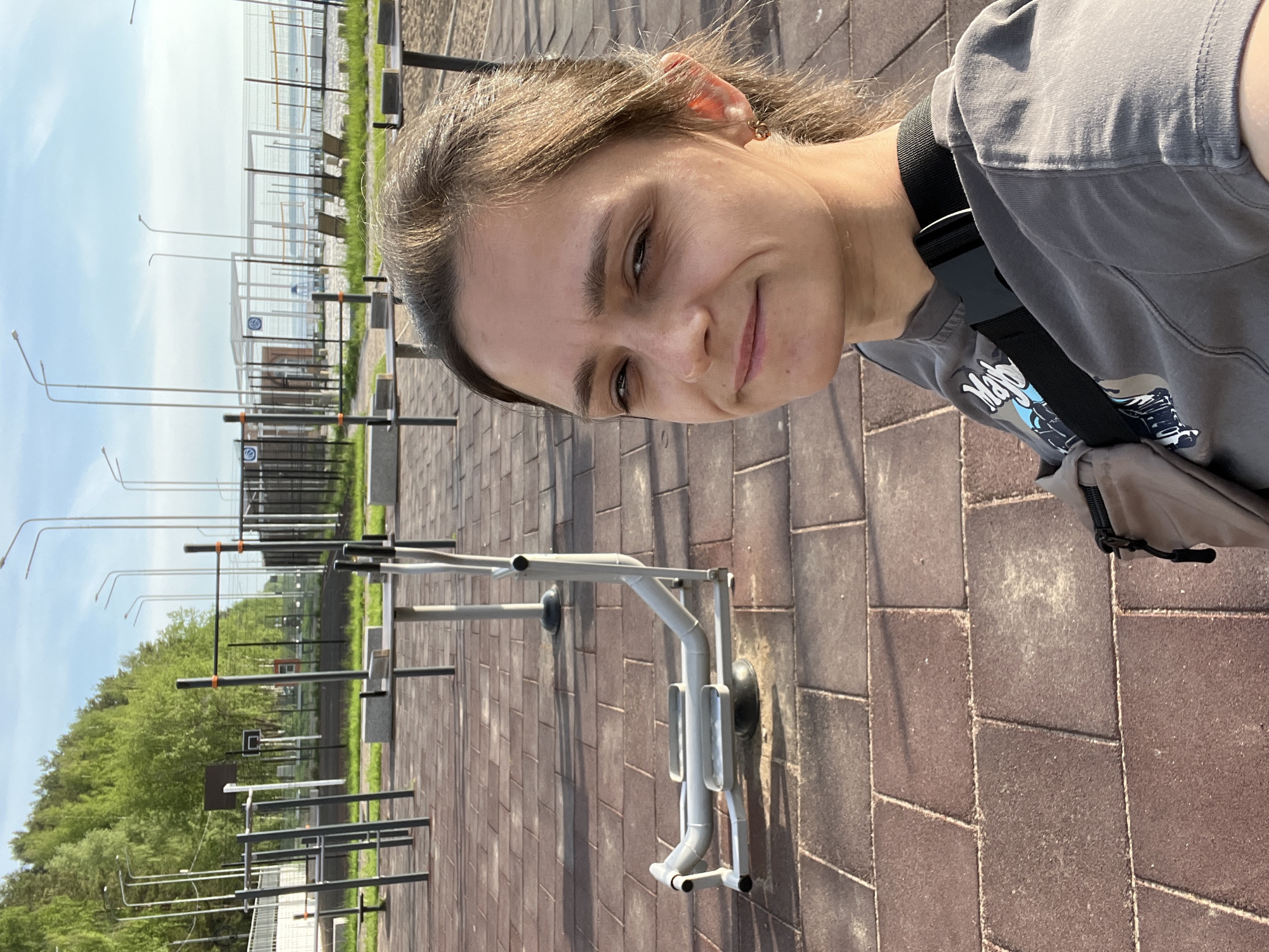 The image shows a selfie of a woman in an outdoor workout area. The location appears to be the city of Chaikovsky, Russia, based on the presence of distinct outdoor exercise equipment that is consistent with photos of workout areas in public parks or squares. The woman is wearing a gray t-shirt and a shoulder bag. The background features paved ground, outdoor exercise equipment, streetlights, a fence, trees, and a blue sky.