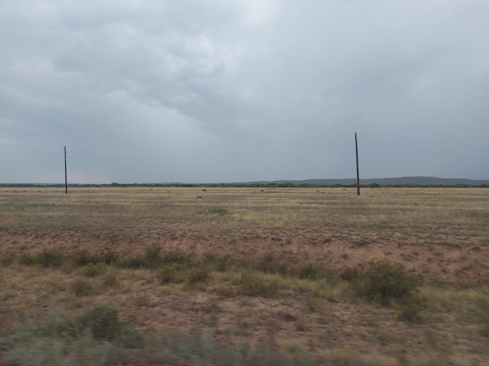 The image depicts a vast, open grassland under a cloudy sky. Two utility poles with wires stretch across the horizon, suggesting a rural or undeveloped area. The ground is covered in dry, yellowish-brown grass and scattered patches of reddish-brown soil. In the distance, a line of low, rolling hills can be seen. The lighting suggests it may be overcast or approaching dusk. The scene is devoid of any human presence or discernible activity. The location is described as Ақтас, Kazakhstan.