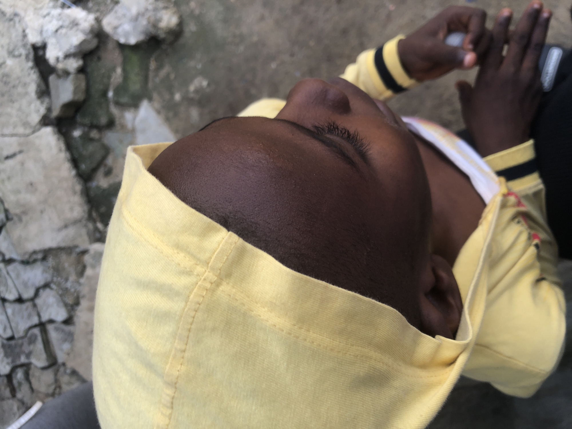 The image shows a close-up, high-angle view of a young child wearing a yellow hooded jacket with black and yellow striped cuffs. The child's face is dark-skinned and partially obscured by the hood, with visible eyelashes and a portion of the nose. The child's hands are in view, with one hand holding a small, light-colored cylindrical object. The background consists of rough, textured surfaces, including what appears to be concrete or stone and some dirt or water. The scene is likely outdoors.