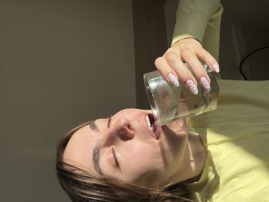 The image displays a close-up of an adult female in an indoor setting. Her eyes are closed, and her mouth is open with the tongue slightly visible, engaged in the act of drinking clear liquid from a transparent, cylindrical glass held in her left hand. Her left hand shows almond-shaped fingernails painted white with a pattern of pink and red speckles. She wears a light yellow long-sleeved garment. Direct, strong sunlight from an unseen source to the right illuminates her face and arm, creating distinct highlights and shadows. The background consists of a plain, light brown-grey wall, possibly indicating an upper floor or attic room with a sloped ceiling. A dark electrical cable is partially visible in the lower right foreground. No identifying visual elements are present to specify a precise location within Izhevsk, Russia.