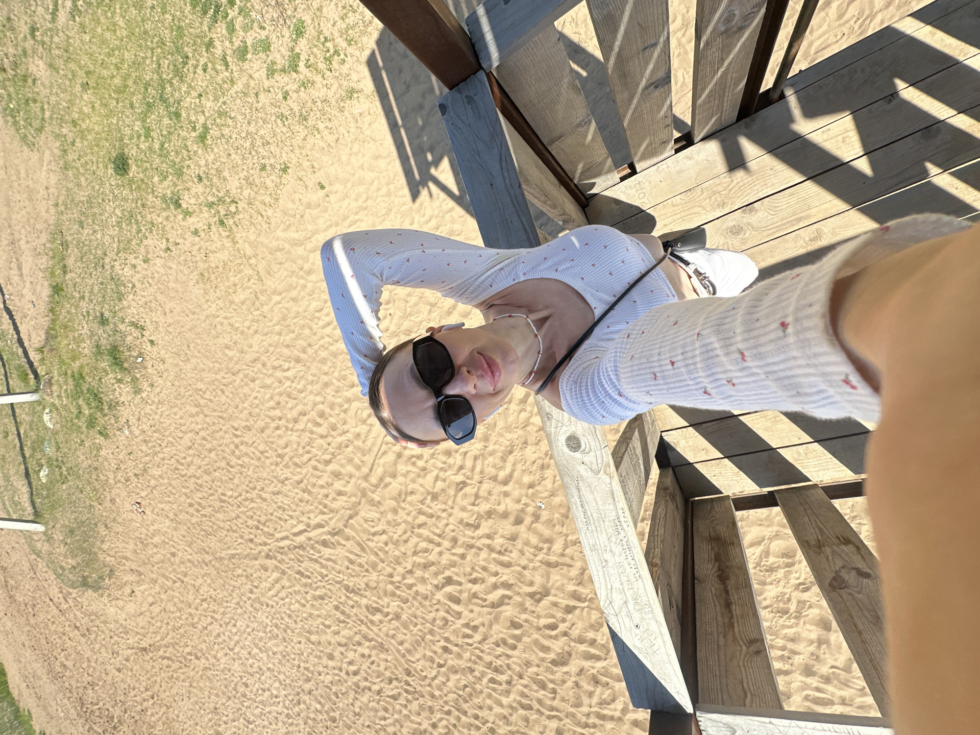 A woman wearing sunglasses, a white long-sleeve top, and a necklace stands on wooden steps, looking up at the camera. The foreground shows wooden steps and railings, casting shadows on the sand. The background features a sandy area with some vegetation. The photo was taken in Votkinsk, Russia.