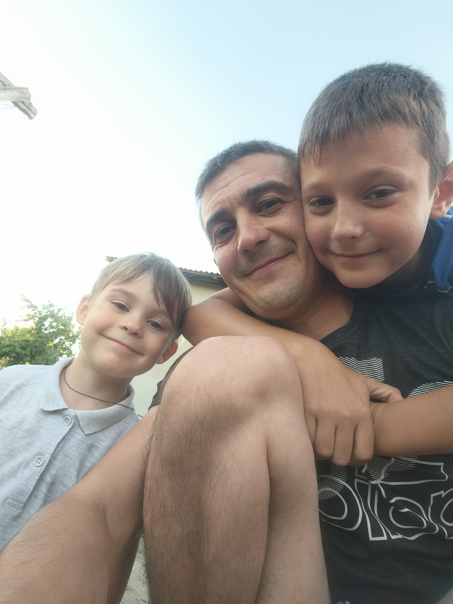 In this candid selfie, a man and two young boys are pictured outdoors. The man, with short dark hair and a slight stubble, is in the center, embracing one boy who is nestled against his chest. This boy has short, light brown hair and is wearing a dark t-shirt. Another boy, with lighter brown hair and a gentle smile, is positioned to the left, looking towards the camera. He is wearing a light grey polo shirt with buttons.
The setting appears to be a backyard or outdoor area, with a hint of a house roof and trees in the background. The sky above is bright and clear, suggesting it is daytime, likely late afternoon or early evening, given the soft lighting. The overall mood is warm and affectionate, with the subjects smiling and close together, indicating a strong familial bond. There is no visible text in the image. The image context places the location in Grinăuți, Moldova.