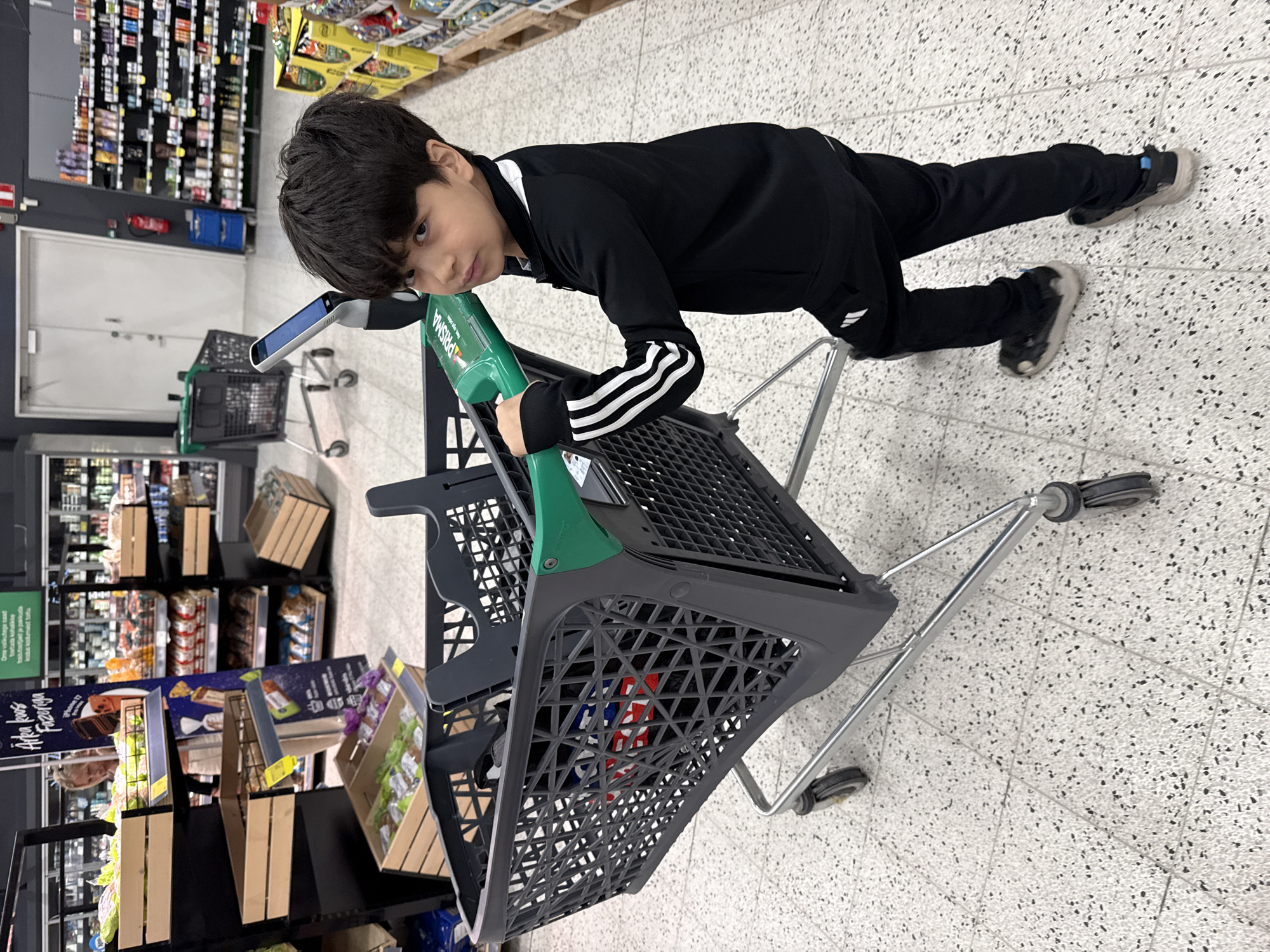 A young boy, appearing to be of Middle Eastern descent, stands in a brightly lit supermarket aisle, pushing a large gray shopping cart. He is wearing a black Adidas tracksuit with white stripes on the sleeves and pants, and dark sneakers. The boy is holding onto the green handle of the shopping cart, which has a blue screen attached to it. Inside the cart, partially visible items include what appears to be a red, white, and blue bag. The background shows shelves stocked with various products, typical of a grocery store. The floor is tiled with a speckled pattern. The setting is identified as a Prisma supermarket in Tallinn, Estonia, based on the provided context.