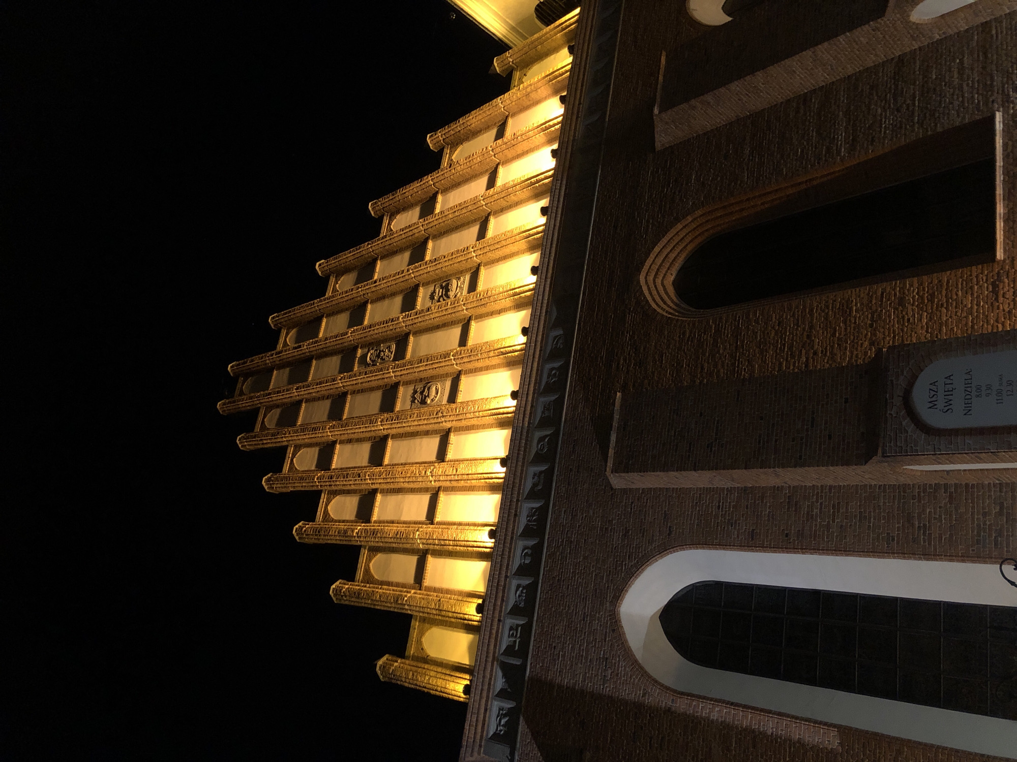An exterior, low-angle night shot features a large brick building, possibly a church, illuminated by warm yellow lights from the top and sides, casting shadows across its facade. The building's architectural style is Gothic, characterized by pointed arches, ribbed vaulting, and ornate detailing along the upper sections. The sky is a deep, inky black, indicating nighttime.
On the lower left of the image, a tall, arched window with a dark pane is visible. To the right of this window, a plaque is mounted on the brick wall. The plaque, written in Polish, reads "MSZA ŚWIĘTA NIEDZIELA 8.00 9.30 11.00 SLOW A 12.30". This indicates the times for Mass on Sundays. The plaque is framed by an arched opening in the brickwork. The building's brickwork is visible throughout the lower portion of the image, showing its textured surface and construction. The upper part of the building displays a stepped facade with vertical elements and decorative reliefs. The overall impression is of a historic and imposing structure at night. Based on the architectural style and the Polish text on the plaque, this building is likely St. John's Archcathedral in Warsaw, Poland.