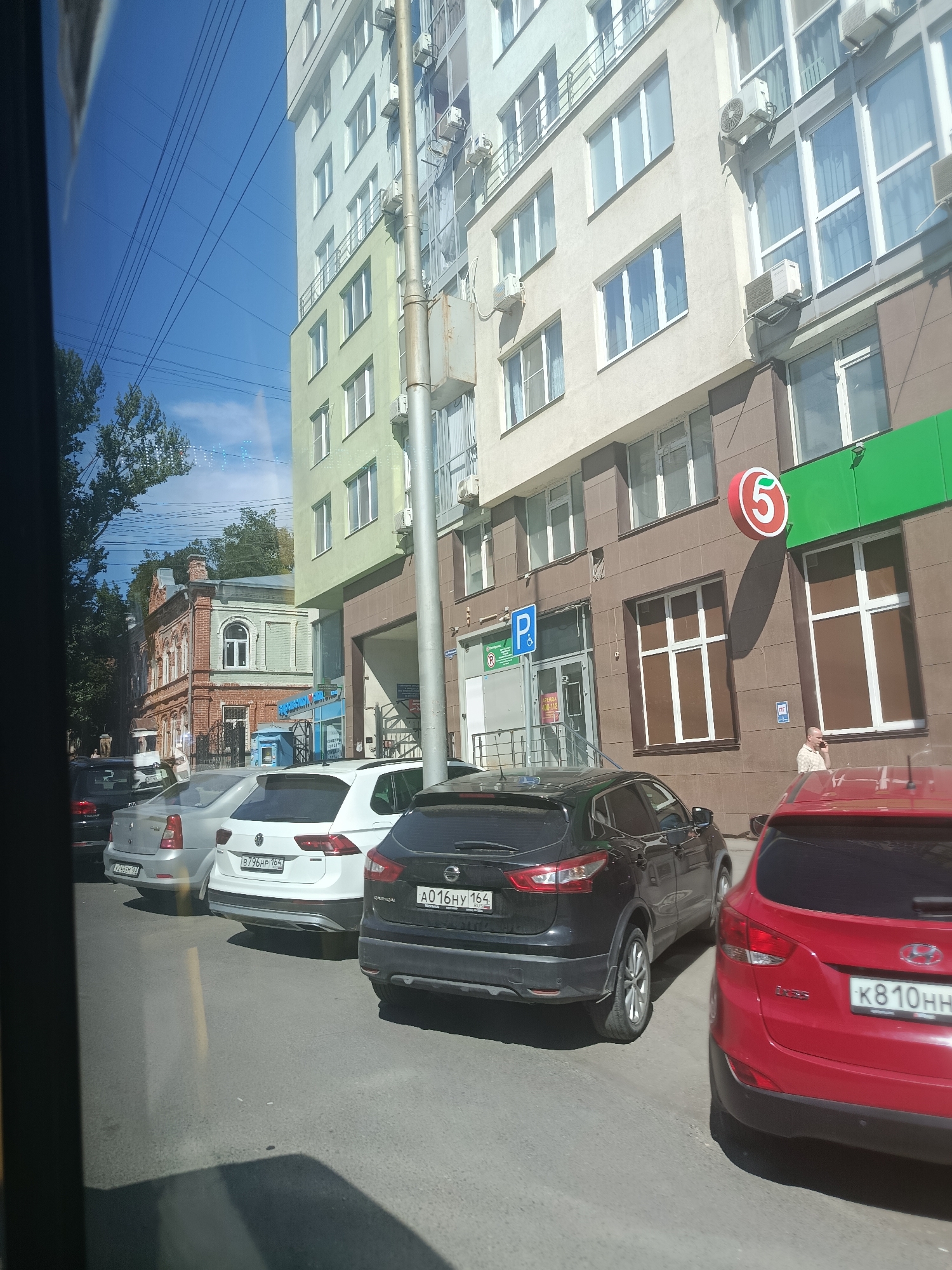 The image captures a street scene in Saratov, Russia, dominated by a modern multi-story building with beige and light green facades. Several parked cars, including a white Volkswagen, a black Nissan Qashqai, and a red Hyundai, line the asphalt road. The building's ground floor features a green facade with a prominent circular red sign bearing the white number "5," indicating a "Pyaterochka" grocery store. A person is visible talking on a mobile phone near the entrance of the building. Above the ground floor, multiple windows and air conditioning units are visible on the building's facade. In the background, an older brick building with ornate architectural details adds a contrasting historical element to the urban landscape. Power lines crisscross the bright blue sky, and the dappled sunlight suggests a clear day.