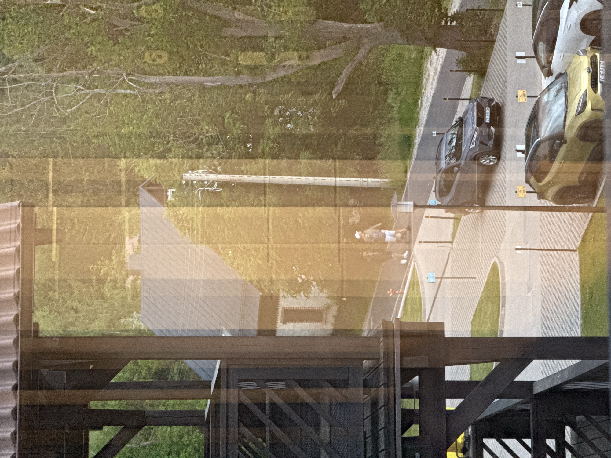 The image depicts a scene in Szklarska Poręba, Poland, as viewed through a window with reflections. In the foreground, a dark wooden structure, likely part of a building's exterior, is visible on the left. Beyond this structure, a paved road curves through a green, wooded area. Two individuals are seen walking on the paved area. One is wearing a white cap and light-colored shorts, while the other is wearing dark clothing. A red fire hydrant is situated on the side of the road.
Further down the road, parked cars are visible. A dark gray SUV with license plate "ZS 341SW" is parked on the right side of the road. In front of it, a yellow SUV with license plate "DJF PY37" is parked. A white car is partially visible to the right of the yellow SUV. Several parking markers with numbers are present. The background is filled with lush green trees, indicating a natural, possibly mountainous, environment characteristic of Szklarska Poręba. Overhead, the sky appears bright, suggesting daylight. The overall scene suggests a quiet, residential or resort-like area with a blend of architecture and nature.