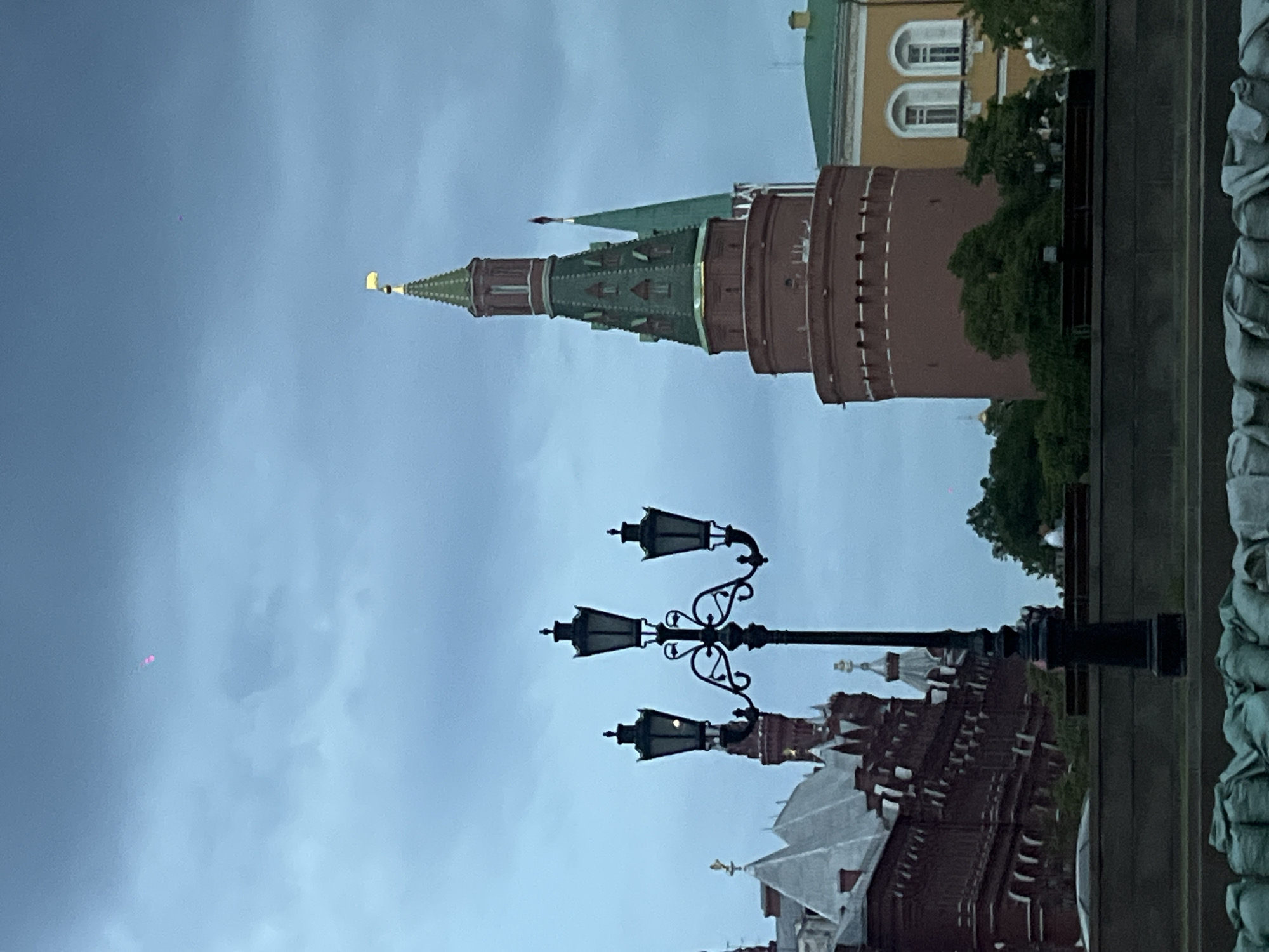 The image shows a view of the Moscow Kremlin in Russia. The focus is on one of the Kremlin towers, characterized by its red brick construction and a green, pointed roof topped with a golden star. A street lamp with three ornate lanterns stands in the foreground. In the background, parts of other Kremlin buildings are visible. The sky is overcast, suggesting a cloudy day. The image is taken at ground level, giving an upward perspective of the tower and the sky.