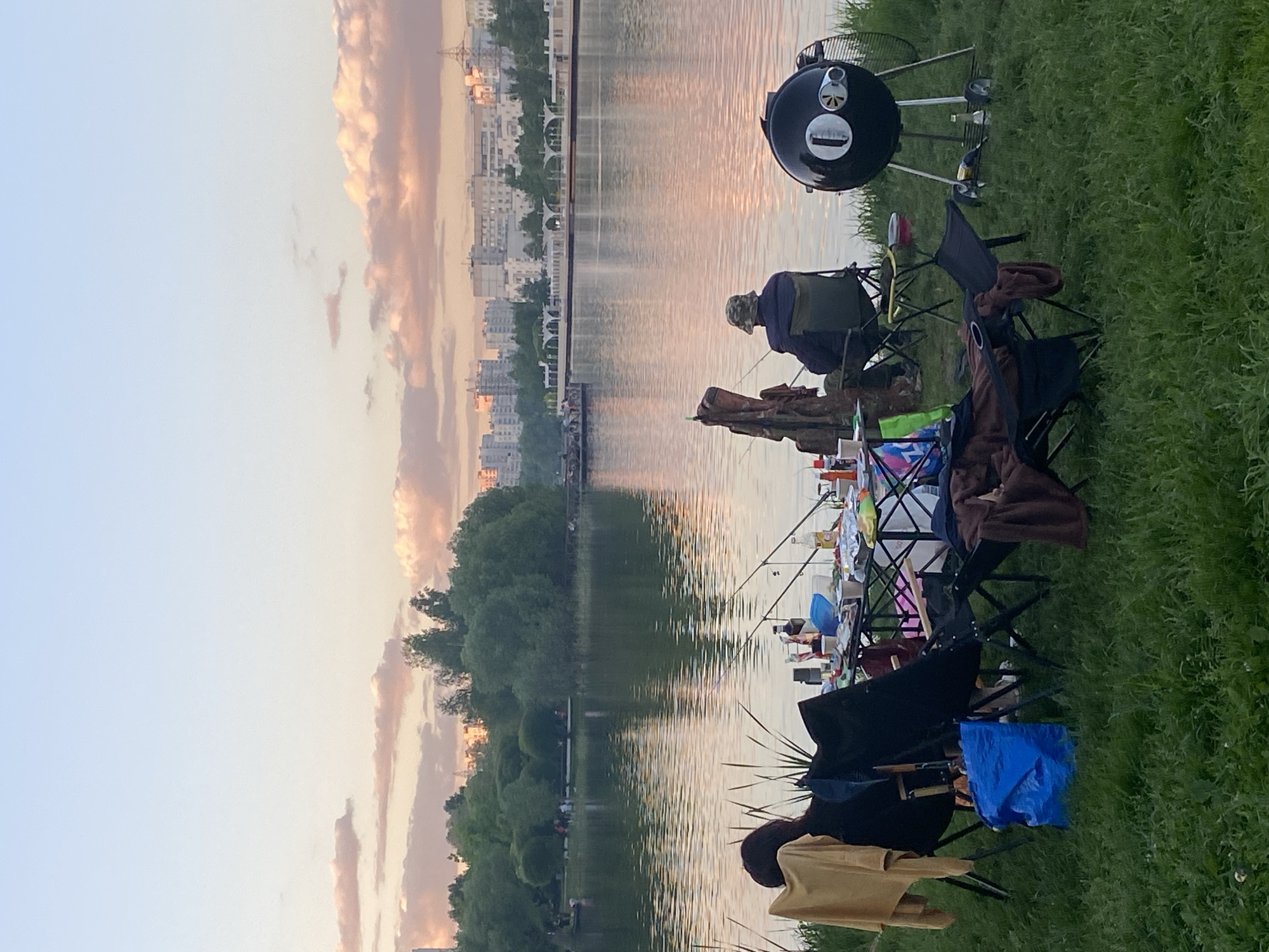 The image shows a group of people fishing and grilling by a body of water, likely a lake or river, in Moscow, Russia. Several fishing rods are cast into the water, and folding chairs are set up along the grassy bank. A grill is visible to the right. In the background, a cityscape is silhouetted against the sky, with clouds reflecting the colors of the sunset. The people present are engaged in fishing and appear to be relaxing by the water.