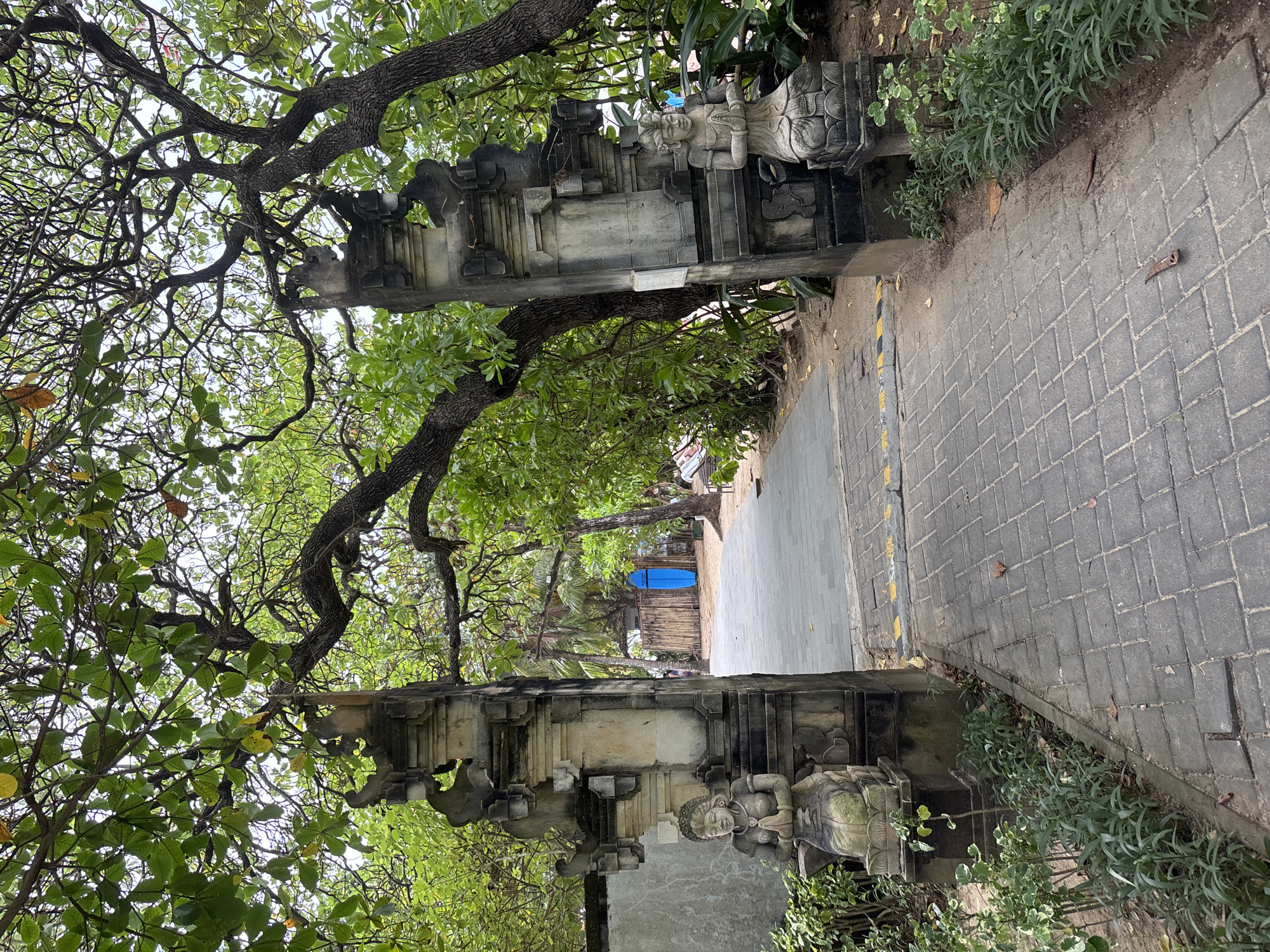 An outdoor pathway made of grey cobblestones leads through a decorative stone archway in Kuta Selatan, Indonesia. The archway features intricate carvings and two stone statues of figures with hands pressed together in a prayer-like gesture, one on each side. Lush green trees with dense foliage arch over the pathway, casting shadows. Beyond the archway, a sandy area is visible with a bamboo fence and a bright blue surfboard standing upright. The environment appears to be a tropical, possibly beachside, location, with vegetation and sand suggesting a warm climate. The image captures a serene and culturally rich entrance.