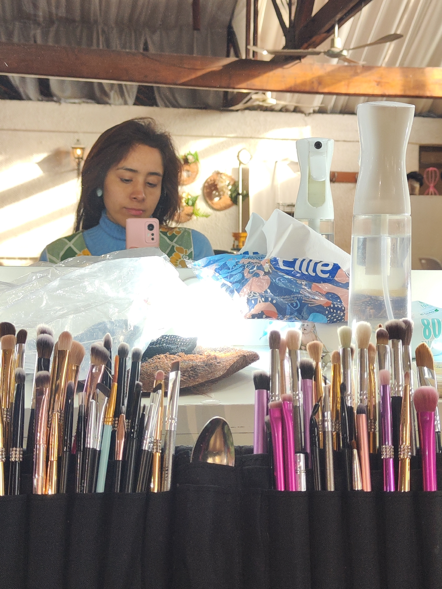 The image shows a reflection in a mirror of a young woman holding a pink smartphone, taking a selfie. She is wearing a blue turtleneck sweater with a green and yellow patterned vest. In the foreground, a black makeup brush organizer is filled with numerous makeup brushes of various sizes and colors, many with metallic handles. Behind the brushes, there is a pile of what appears to be a brown, textured object, possibly a decorative item or a piece of furniture, and a plastic bag. To the right of the organizer, there are two white spray bottles and a package of facial tissues with colorful branding. The background of the reflection shows a ceiling with wooden beams and a ceiling fan, as well as some decorative wall hangings. The lighting suggests natural sunlight streaming in. This scene is likely from a residential or studio space in Santiago, Chile.