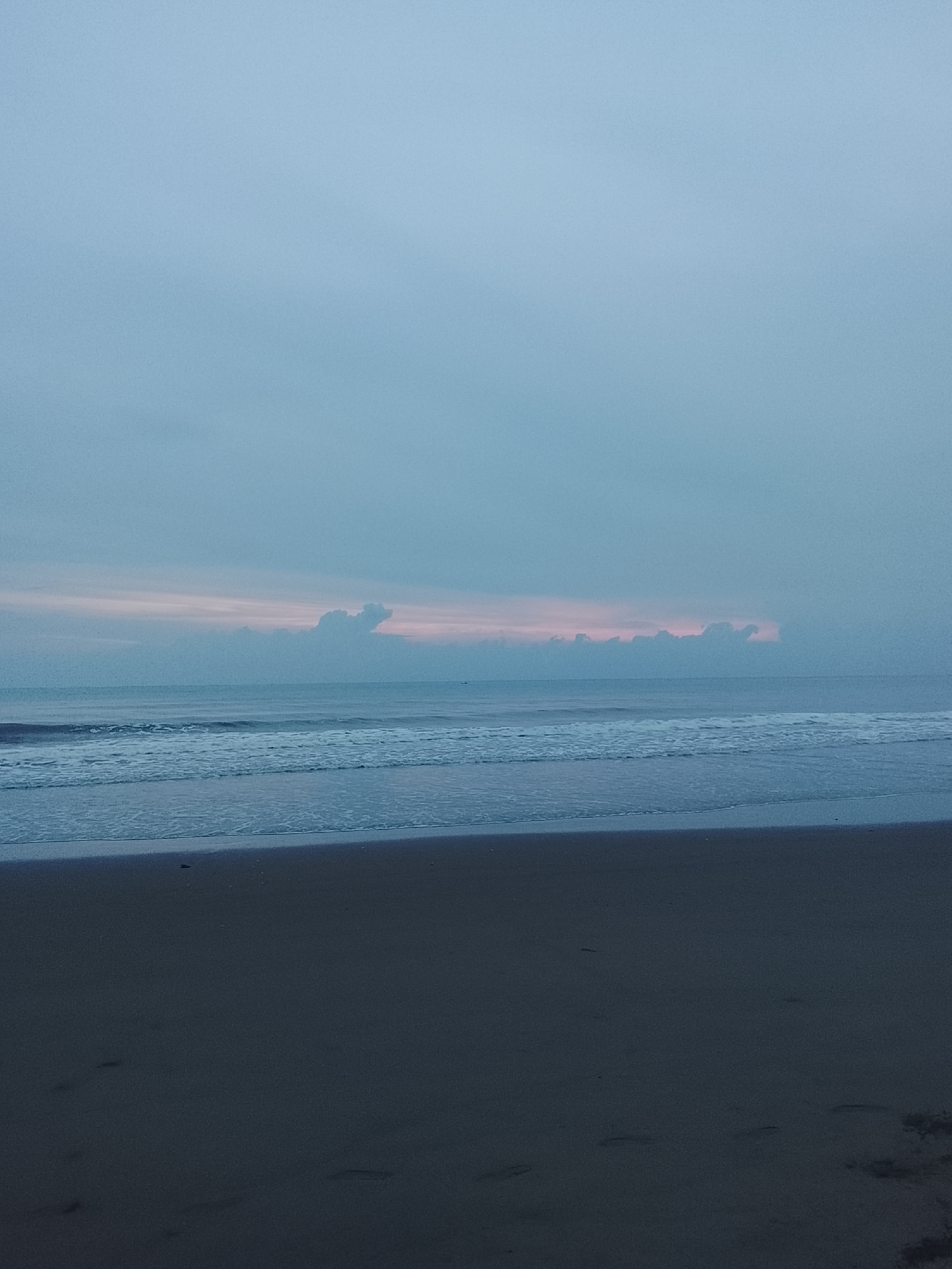 A wide shot captures the tranquil beach of Reudeuep, Indonesia, under a soft, overcast sky. The ocean stretches out to the horizon, with gentle waves rolling onto the dark sand shore. A faint pinkish hue is visible on the horizon line, hinting at either sunrise or sunset, partially obscured by low-lying clouds. The foreground is dominated by the dark, damp sand of the beach, with a few indistinct footprints scattered across its surface. The overall atmosphere is serene and peaceful, with no visible people or man-made structures, emphasizing the natural beauty of the coastal landscape.