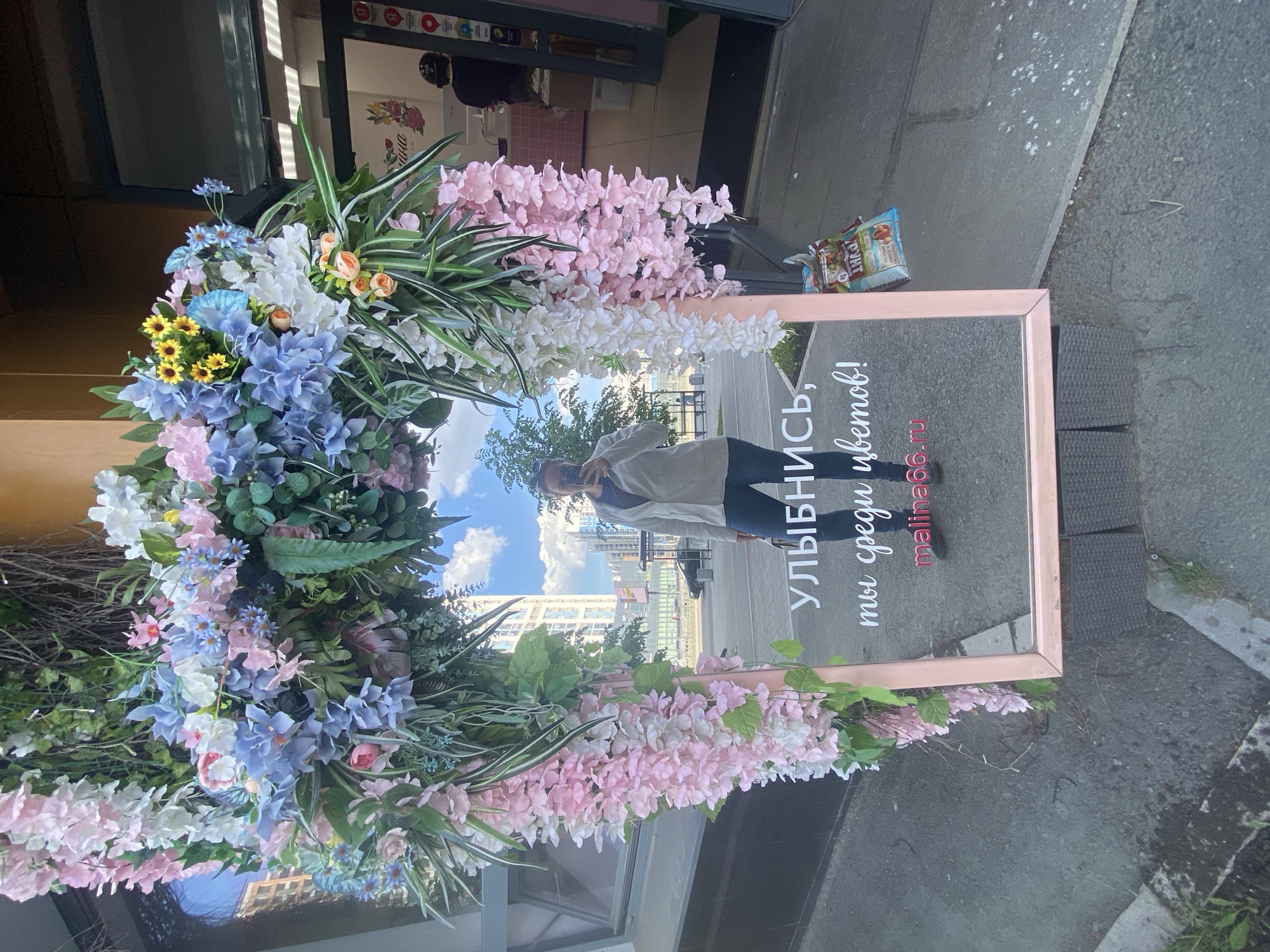 The image depicts a woman's reflection in a framed mirror decorated with artificial flowers. The mirror is set up on a city sidewalk in Yekaterinburg, Russia. The floral arrangement around the mirror includes a mix of pastel-colored flowers, particularly pink and blue hydrangeas, white orchids, and greenery. The mirror's frame is a light pink. Reflected in the mirror is the woman taking the photograph, a partial view of a city skyline, and the sky. The text on the lower portion of the mirror reads "Улыбнись, ты среди цветов! malina66.ru". To the side of the mirror, on the ground, there is a discarded snack bag. The sidewalk is concrete.