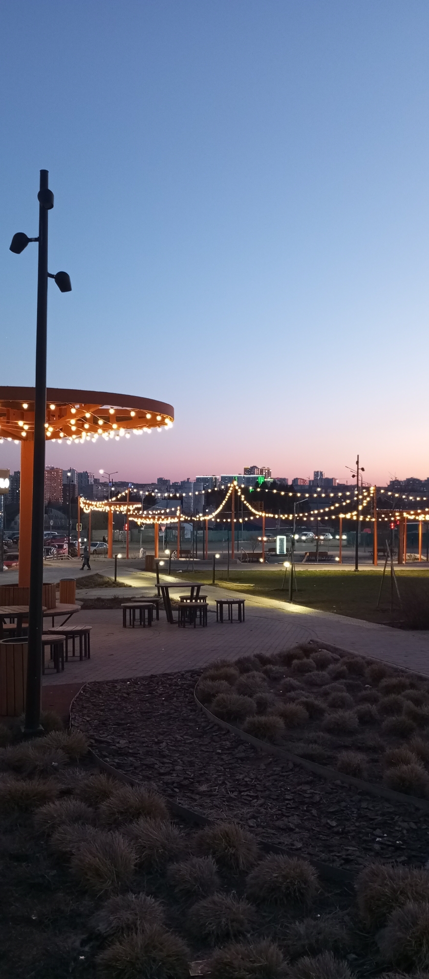 This is an outdoor evening scene in Pervomaysky, Russia. The sky transitions from blue to a pinkish-orange hue at the horizon.
Illuminated string lights are strung between wooden pergola-like structures that are set up along a brick pathway. There are several picnic table sets along the pathway. The foreground shows a low-lying ground cover separated from the pathway by a metal border. A tall streetlamp with multiple light fixtures is on the left. In the background, there are apartment buildings. A lone figure can be seen walking along the lit pathway. A few cars are visible in the background as well.