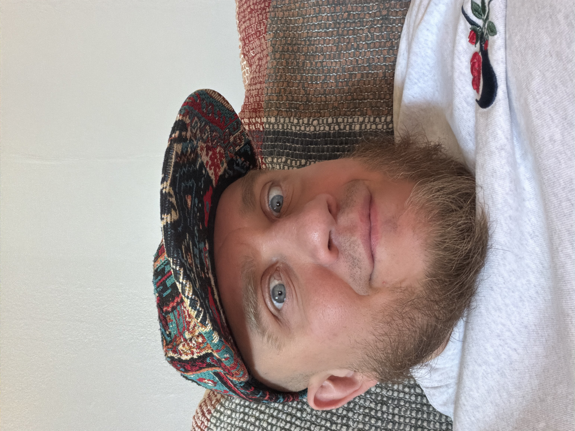 A man with light skin, blue eyes, and a beard is looking directly at the camera. He is wearing a colorful patterned hat and a white shirt with an embroidered flower design near the collar. The background includes a textured wall and a patterned cushion. The location is unspecified in the city of Morton Grove, United States of America.