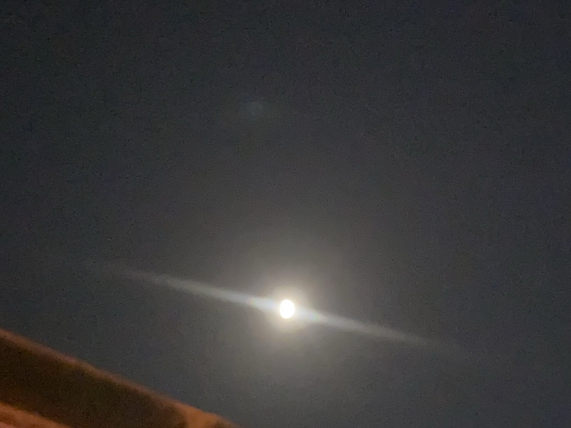 The image shows a night sky with the moon as the primary subject. The moon is a bright, luminous sphere in the center of the frame, emitting a strong light that creates a visible lens flare or halo effect. A beam of light extends upwards from the moon, suggesting a diffusion of light through atmospheric conditions or an optical artifact. In the upper left corner, a portion of a dark, textured object, possibly a building or a natural element like a tree branch, obstructs a small part of the sky. The background is a deep, dark gray, indicating a nighttime setting. The overall scene conveys a sense of nighttime ambiance in an outdoor environment. The location in Sabon Gari West, Nigeria, places this scene within a specific urban or peri-urban context at night.