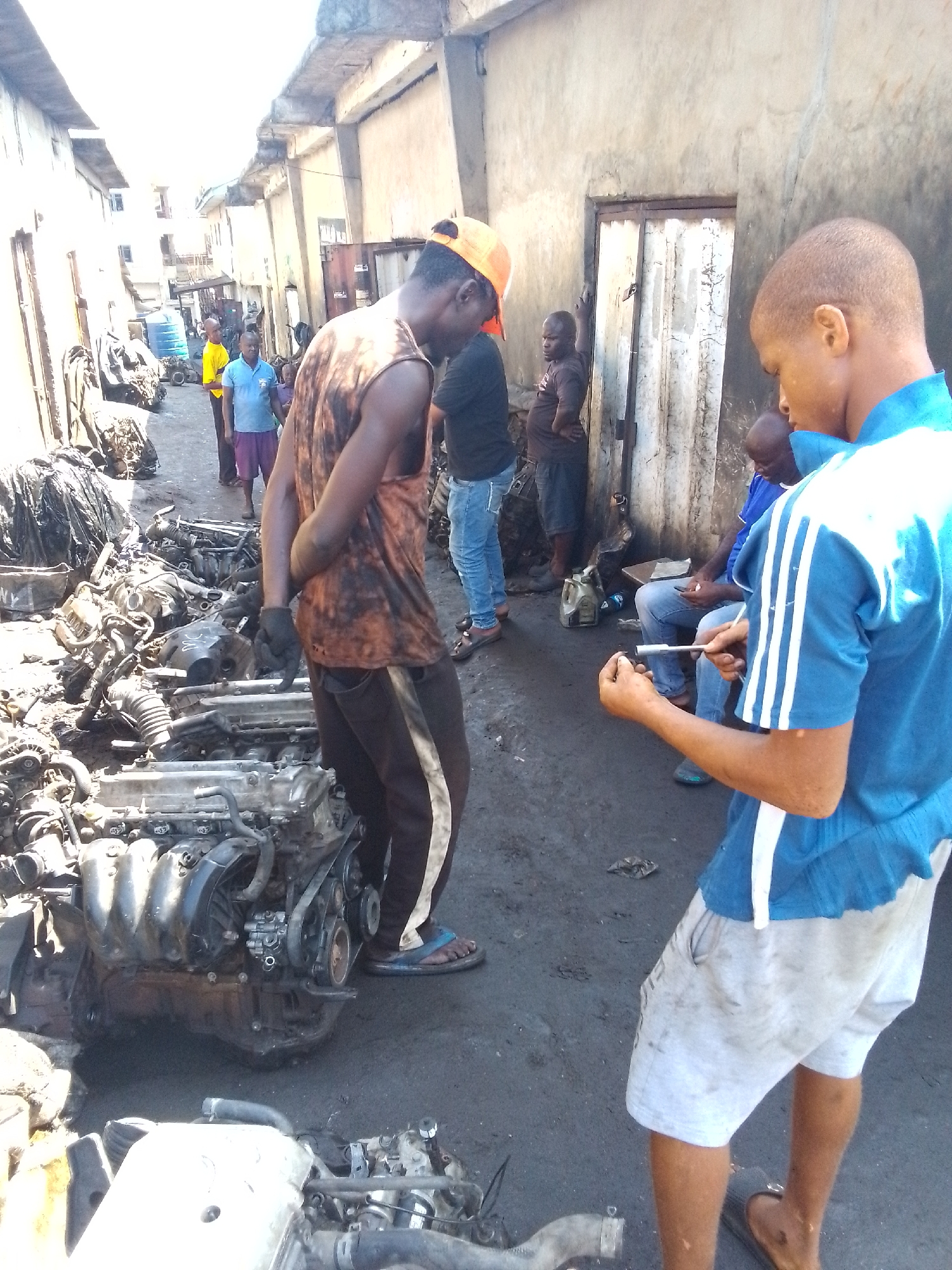 The image captures a scene in what appears to be an auto parts market in Onitsha, Nigeria. The environment suggests a busy, informal business area, with buildings lining a narrow street. Disassembled engines and auto parts are scattered on the ground, indicating the primary activity is the buying, selling, and repair of automotive components.
Several individuals are visible. One man in the foreground is wearing a blue shirt with white stripes on the sleeves and light-colored shorts. He's holding a small part and a tool, possibly inspecting or working on the part. Another man, facing away from the camera, is wearing an orange sleeveless shirt, dark pants with a white stripe, and a cap; he has his hands clasped behind his back. Several other individuals are standing or sitting in the background.
The light suggests it's daytime, possibly midday, in a well-lit outdoor location. The overall impression is that of a vibrant, if somewhat cluttered, marketplace, with a focus on automotive parts and repair.