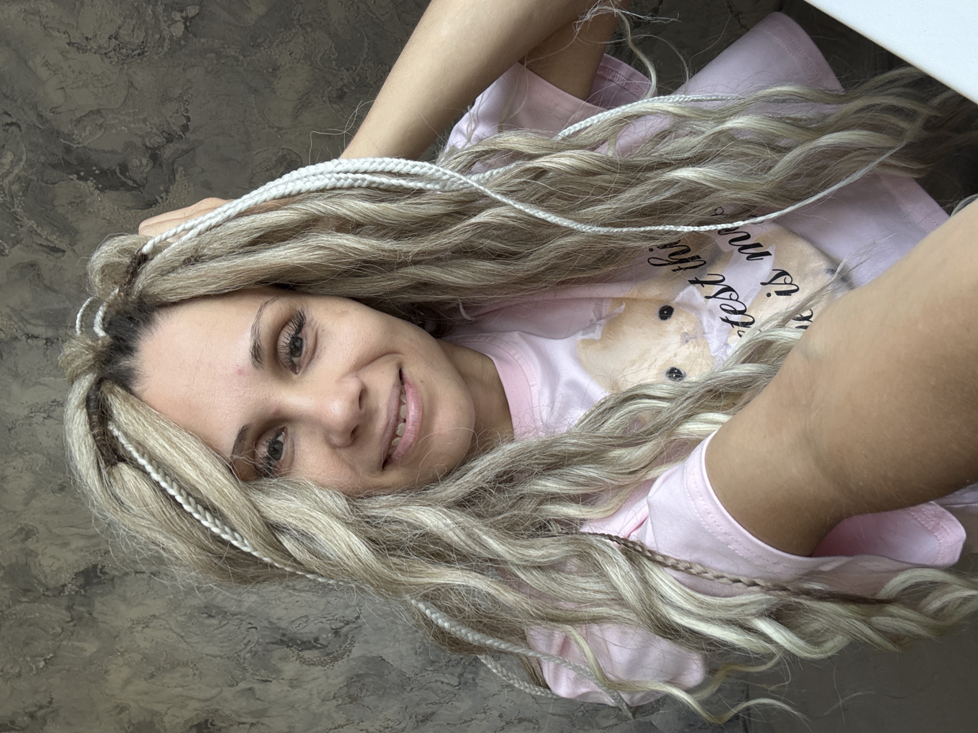 The image is a high-angle close-up of a woman indoors. The woman has long, light blonde hair that appears to be styled in braids or dreadlocks, with some sections incorporating white or brown accent braids. She is wearing a light pink t-shirt. The background is a grey textured surface, possibly a floor or wall. The setting does not appear to be in Rusanovo, Russia, based on the details available within the image.