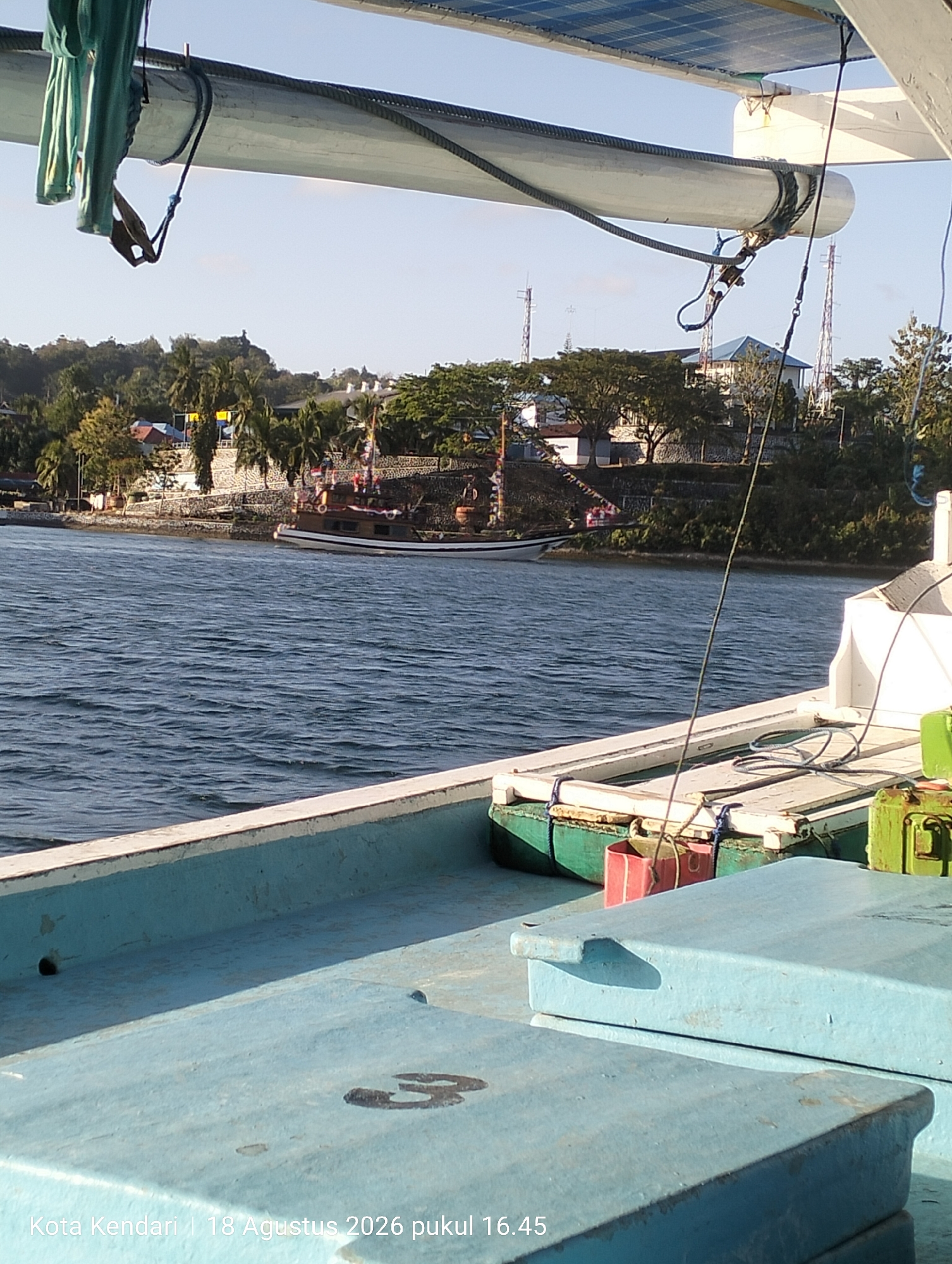 A wide-angle shot captures a view from the deck of a boat on a body of water. In the foreground, light blue hatches and some green and red containers are visible on the boat's deck. In the midground, the rippling water of a bay or strait is seen, with a distant shore line featuring a stone wall, vegetation, and buildings. Several small boats, including a larger wooden vessel, are docked or sailing near the shore. Tall communication towers are also visible on the land. The sky above is clear and bright. Text at the bottom of the image reads "Kota Kendari | 18 Agustus 2026 pukul 16.45," indicating the location as Kendari, Indonesia, and the date and time as August 18, 2026, at 4:45 PM.