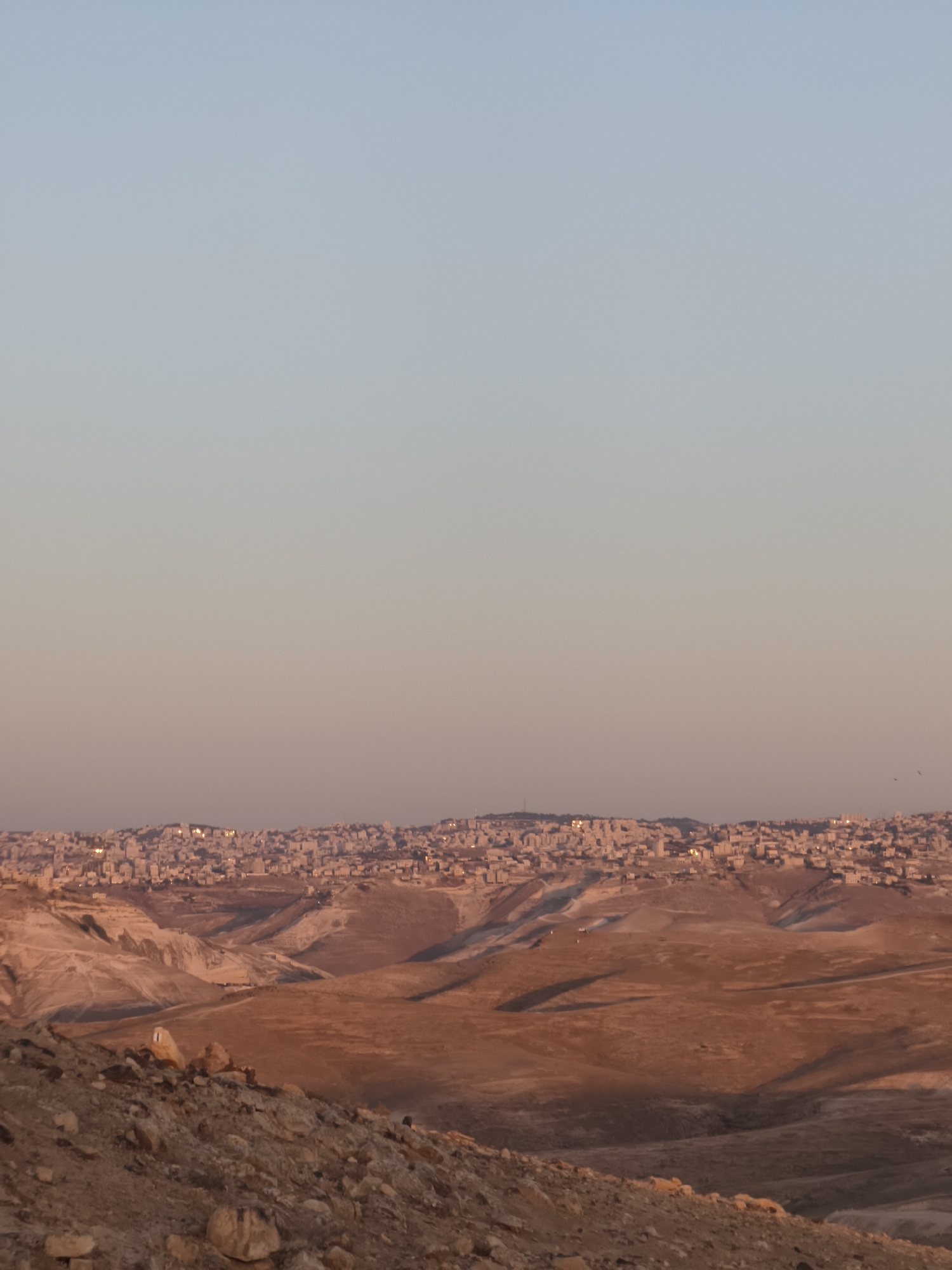 The image depicts a vast, arid landscape under a pale blue sky that transitions to a warm pink hue near the horizon. The foreground is dominated by rocky, barren terrain with scattered boulders and pebbles. In the mid-ground, rolling hills and valleys, characterized by dry earth in shades of brown and ochre, are visible. A distant city, with densely packed buildings and illuminated windows suggesting evening or dusk, sprawls across the horizon. The city appears to be built on a hilly terrain, with structures appearing as small, light-colored dots against the darker landscape. The overall impression is one of a remote, undeveloped area with a burgeoning urban settlement in the distance.