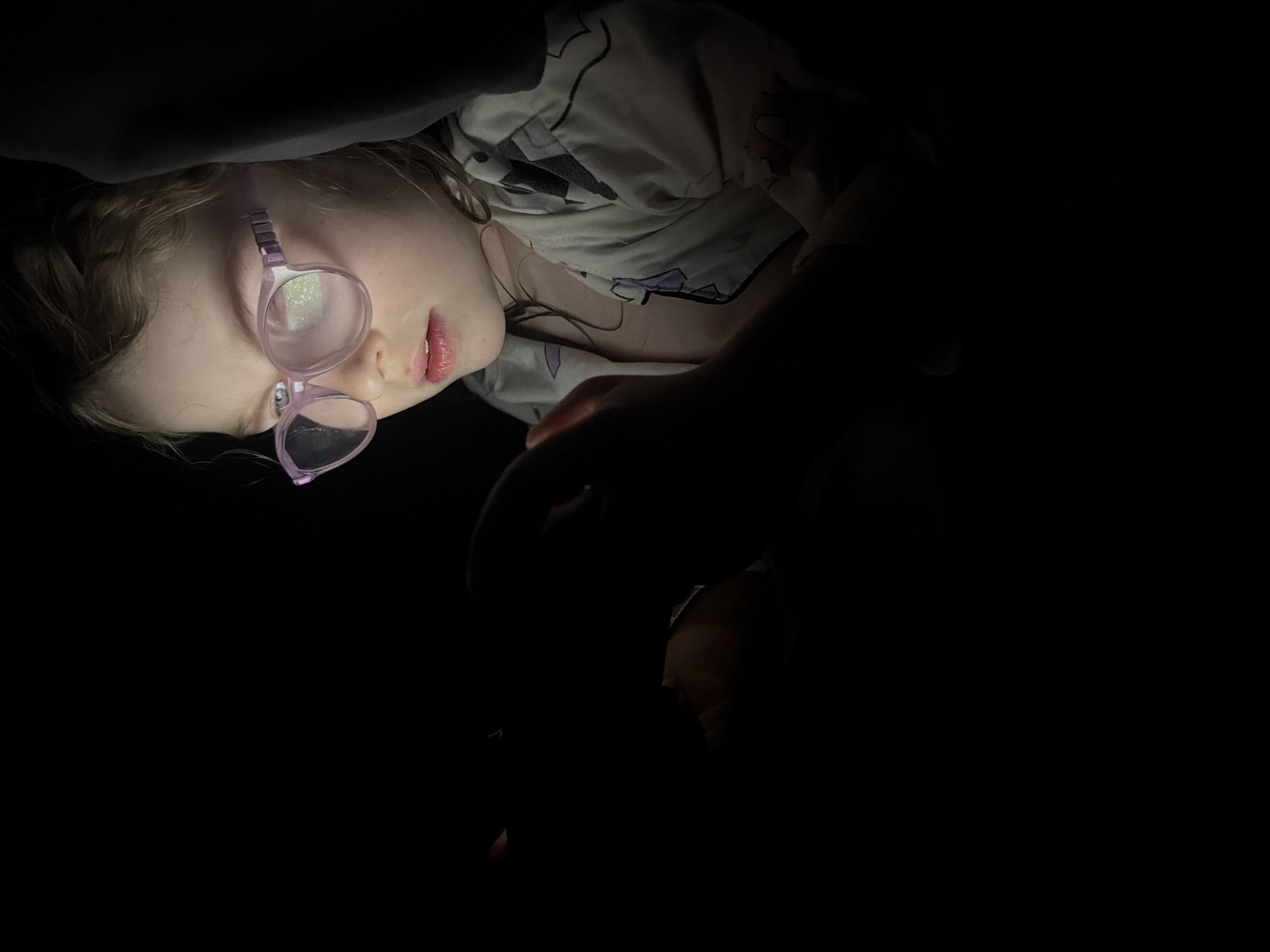 A close-up, dimly lit image shows a young girl lying down, likely indoors, in what appears to be a dark room or under covers. Her head is tilted, and she is looking upwards and slightly to the right, towards an unseen light source that illuminates her face from below. This strong, localized light creates highlights on her skin and significant reflections on her purple-framed glasses, making it difficult to see her eyes clearly through the lenses.
The girl has fair skin and light brown or blonde hair, partially visible around her face. Her lips are slightly parted, revealing a natural pink hue, and her expression is neutral, suggesting focus or contemplation. She is wearing what appears to be sleepwear or a comfortable top with a subtle light-colored pattern (possibly white with purple or grey shapes). A dark fabric, possibly a blanket or pillow, is visible above and to her left, further emphasizing the dark environment.
The overall impression is one of a quiet, intimate moment, perhaps a child reading or watching something on a device in the dark. The dramatic lighting from below creates a somewhat mysterious or cozy atmosphere. No specific location cues for Chaikovsky, Russia, are visible in the image, nor is any text present. The time of day is clearly night or a very dark setting.