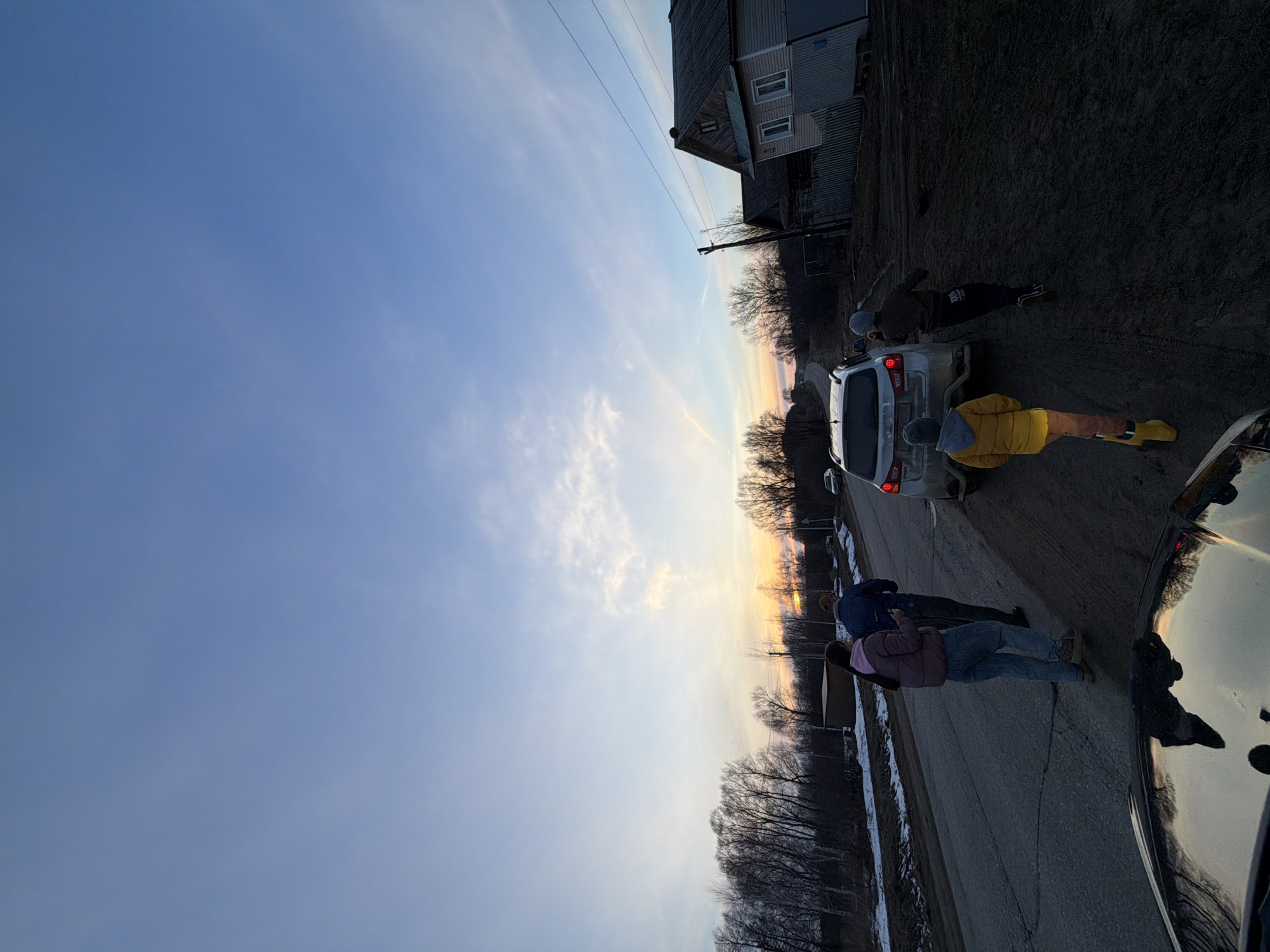 The image depicts a street scene in Дулесово, Russia, at dusk or dawn. The sky is a gradient of blue to orange near the horizon. A two-story building is visible in the background. A white car is parked on the side of the road. Several people are standing near the vehicle. One individual wears a yellow jacket. Another appears to be examining the car's rear. The road has cracks and minor damage. Bare trees line the horizon. Another vehicle's hood is visible in the bottom right corner.