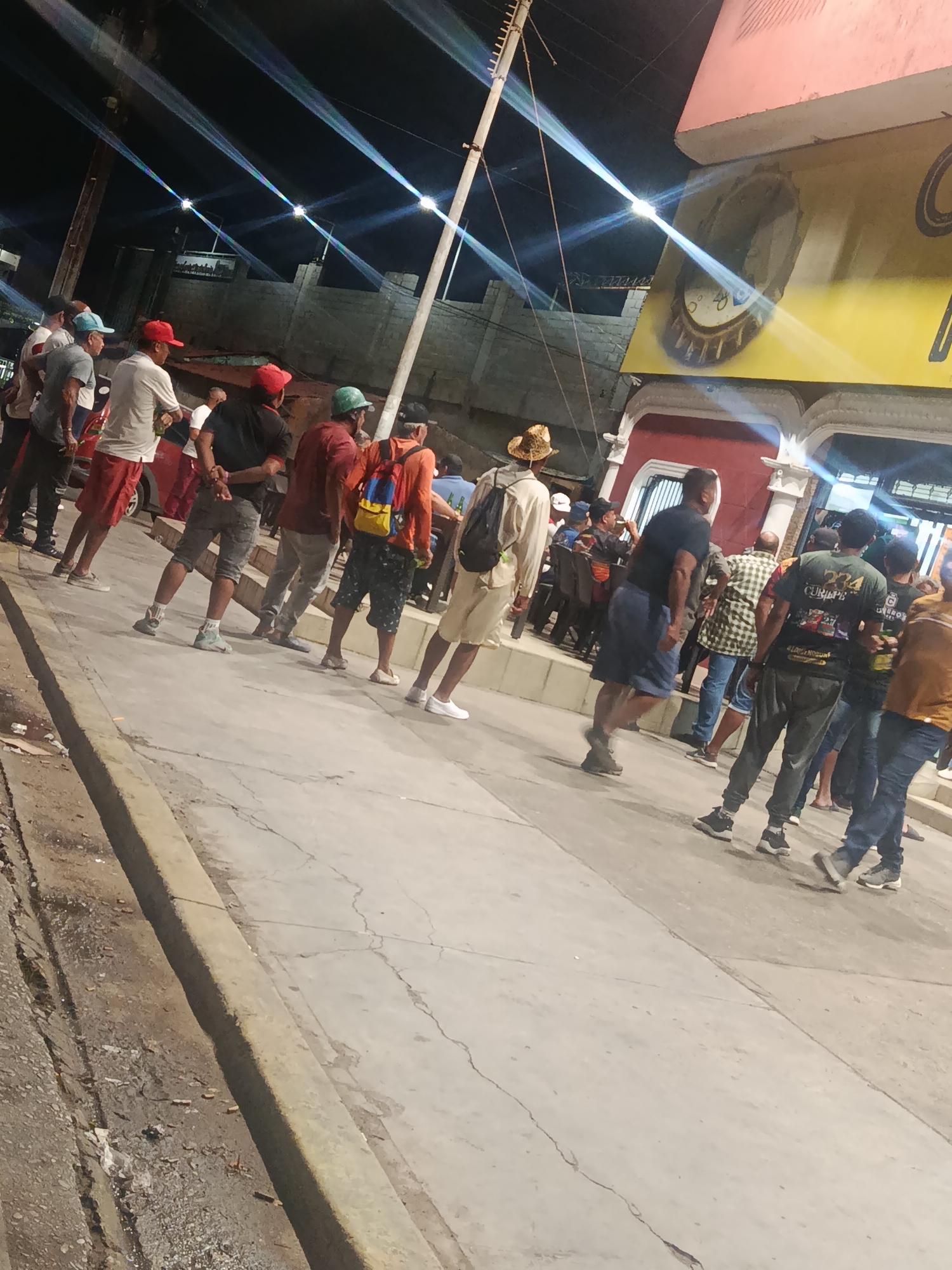 The image depicts a nighttime street scene in Juan Griego, Venezuela, featuring a group of approximately 15-20 adult males gathered on a concrete sidewalk. The sky is dark, illuminated by multiple streetlights creating distinct, bright beams of light.
In the foreground and midground, a concrete sidewalk, showing cracks, extends from the lower left to the upper right. To the immediate left of the sidewalk, a dirt-and-gravel strip contains scattered debris.
On the left side of the image, a line of men faces generally towards the right. Individuals include a man in a light blue cap and grey t-shirt, another in a red cap and white t-shirt with red shorts, and a third in a red cap and black t-shirt with grey shorts. Further back, a man wears a straw hat, light-colored long-sleeve shirt, and shorts, carrying a dark backpack. A red vehicle, possibly a car, is partially visible near this group.
On the right side, another group of men stands or walks. Notable figures include a man in a green cap and orange t-shirt with blue shorts, carrying a blue and yellow backpack, and a man in a black cap and dark t-shirt with patterned shorts. Other individuals in casual attire, predominantly t-shirts, shorts, and sneakers, are visible in varying degrees of clarity.
The environment includes a building on the right with a pink upper facade, a yellow wall below it featuring a large, round, bottle-cap-shaped sign or mural. Beneath this, a red architectural element with white ornate trim is visible. In the background, behind the left group of people, a building constructed of concrete blocks is present. Utility poles are visible on both the left and right sides of the frame. The overall scene suggests a busy, urban gathering at night.