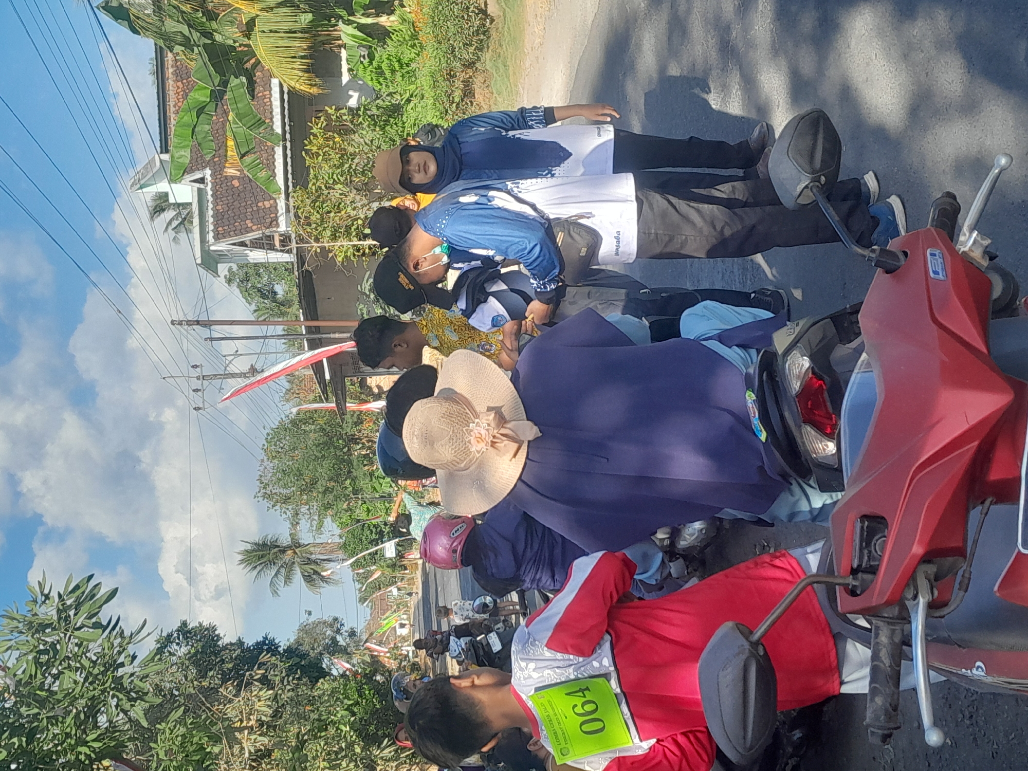 In Sambirejo, Indonesia, a group of people are gathered on a street. A young male in a red and white shirt with the number "064" displayed on his back is facing away from the camera. To his left, a person wearing a purple top and a wide-brimmed hat is seated on a motorcycle. Behind them, several individuals are interacting, including a man in a blue shirt and a woman in a blue headscarf and white shirt. The scene appears to be outdoors, with houses, trees, and power lines visible in the background, suggesting a residential area. The presence of motorcycles indicates transportation is a significant element of the scene. The sky is bright with scattered clouds. Red and white Indonesian flags are visible along the street, possibly indicating a local celebration or event.