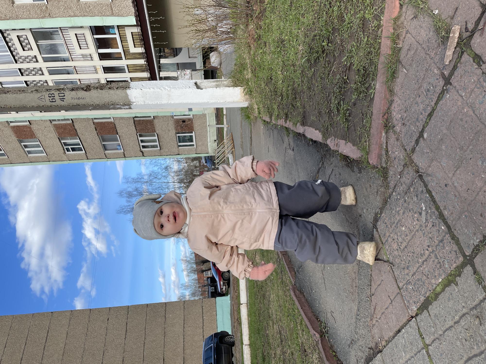 A baby, wearing a grey hat with attached ears, a light pink jacket, grey pants and white shoes, stands on a grey paved sidewalk in what appears to be the city of Izhevsk, Russia. The baby faces the camera with its hands extended to the sides. Behind the baby, there's a patch of green grass and a concrete curb. In the background, there are several concrete apartment buildings, characterized by repetitive window patterns and grey and beige colors. There is also a blue sky with white clouds partially visible. A black SUV car is visible in the background. A concrete structure with the numbers "68 40" painted on it is to the left of the baby.