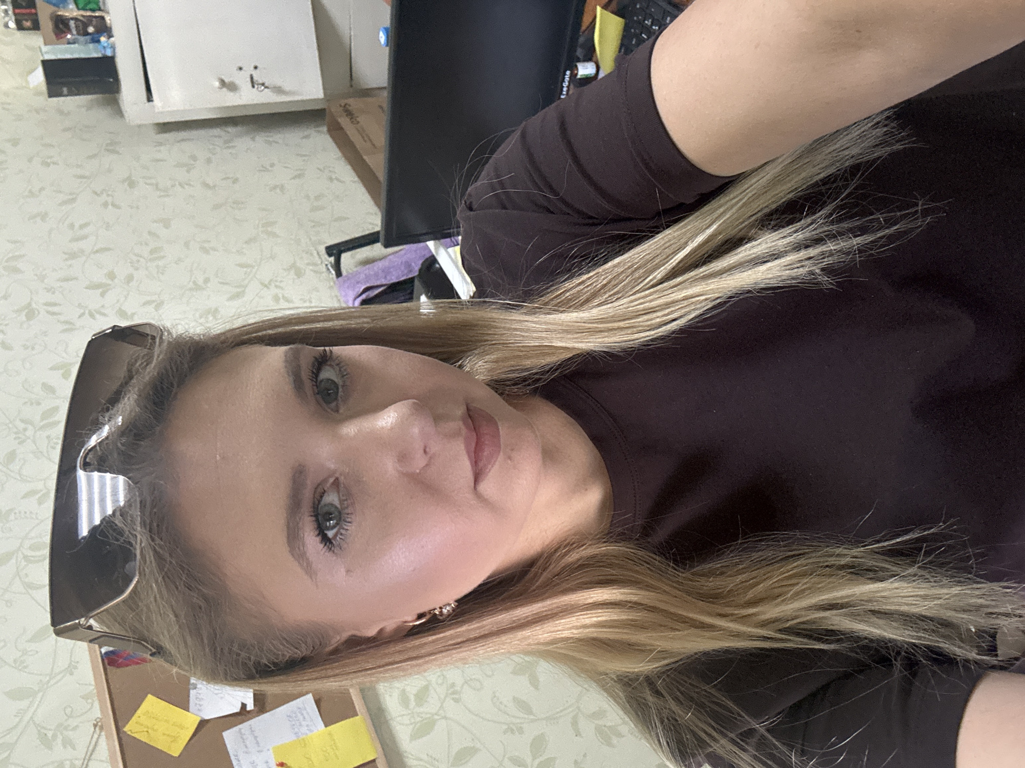 A young woman with blonde hair is taking a selfie in an indoor setting. She is wearing sunglasses on top of her head and a dark brown t-shirt. Her hair is long and falls around her shoulders. In the background, a corkboard with notes is visible, as well as a computer monitor and a white cabinet. The wallpaper has a floral pattern. The lighting suggests it is daytime, possibly in an office or study environment.