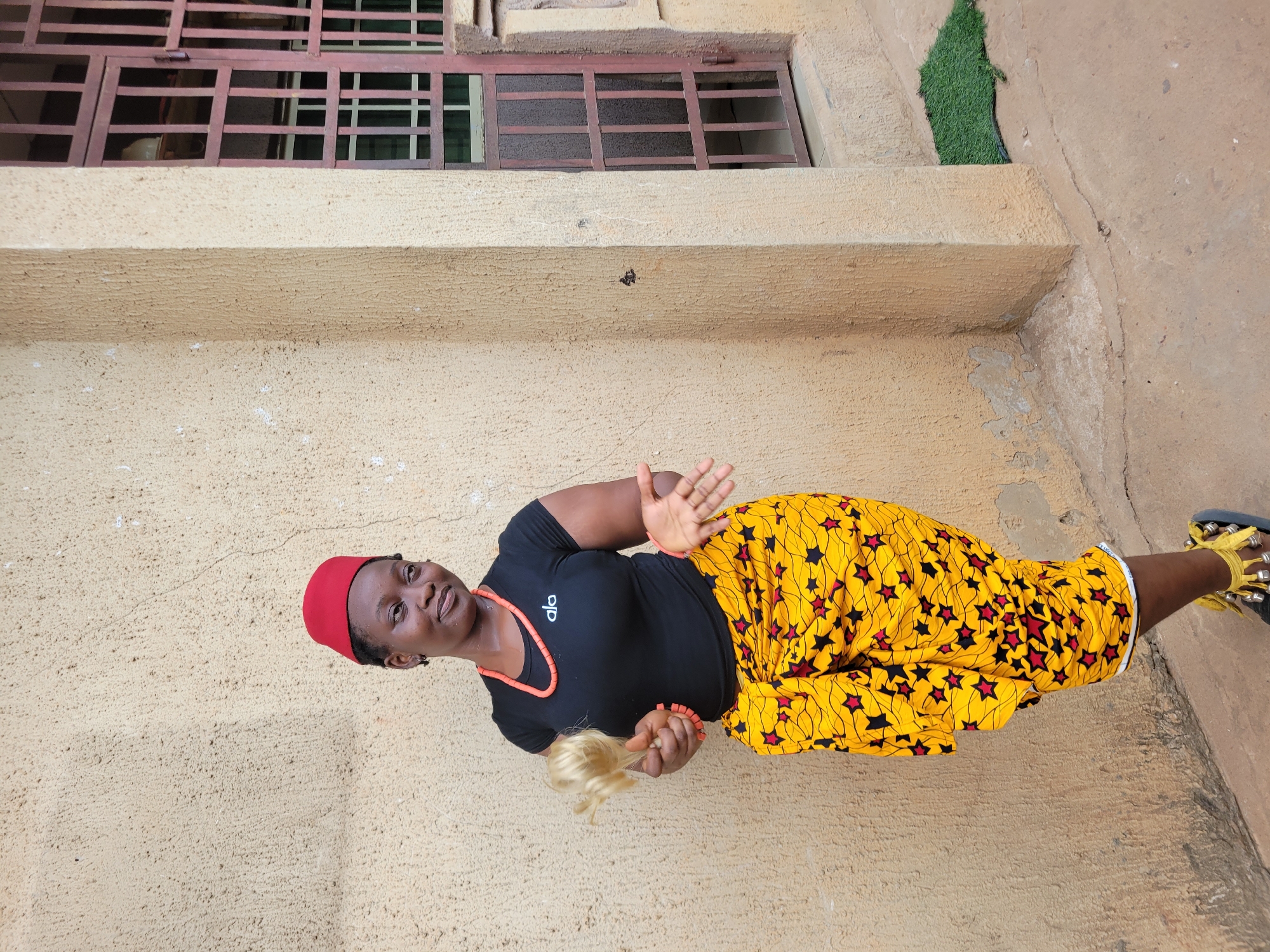 A woman stands outdoors in front of a building. She wears a red cap, a black shirt with an orange neckline, a yellow skirt with red and black star patterns, and yellow sandals with black soles. She has beaded jewelry on her neck and wrist. In her left hand, she holds a blonde-haired object. Her right hand is raised. The building behind her is beige and has a window with a brown metal grate. A patch of artificial green grass is visible above the window. The location is identifiable as Onitsha, Nigeria, based on the woman's attire and jewelry style.
