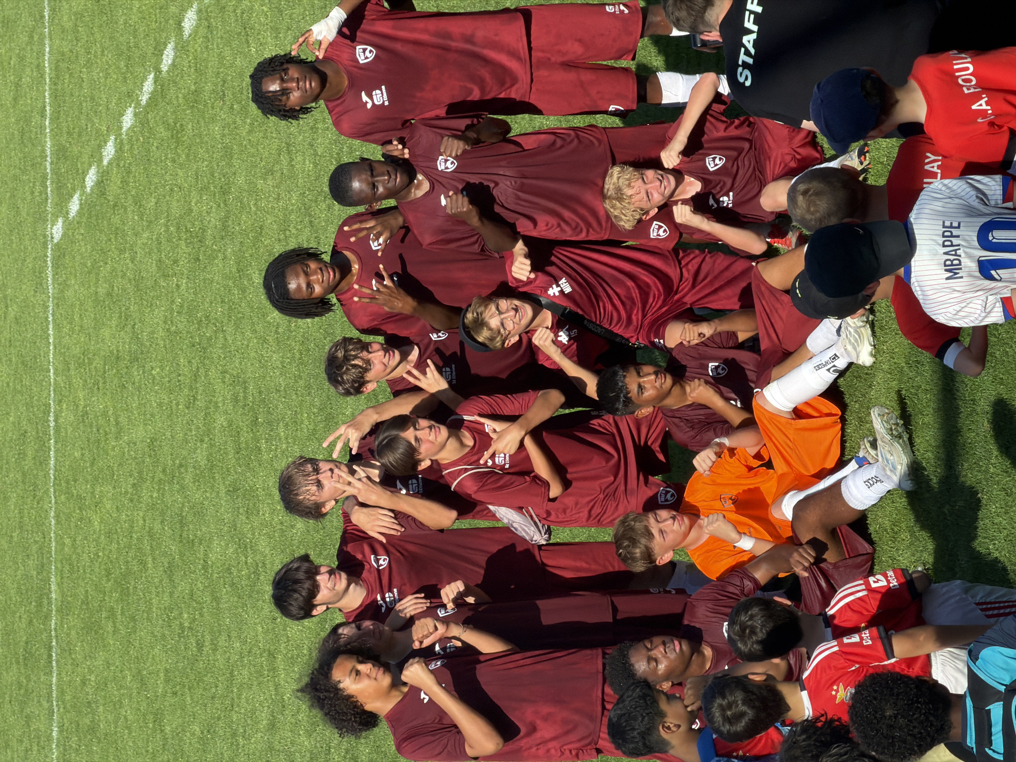 Here is a detailed description of the image:
**General Description:**
The image is an aerial shot of a group of people lying down on a grassy field, likely a soccer field, possibly after a game or training session. The image appears to be taken in bright, sunny conditions.
**People:**
The group consists of about fifteen to twenty people, mostly young males, likely teenagers. Most of them are wearing maroon athletic shirts with a logo that says "SS Etienne" and "MIFA", indicating they are part of a soccer team. The team appears to be diverse in terms of ethnicity. Some are wearing white shorts and socks. One person is wearing an orange shirt. There are others wearing red shirts and some in striped white and blue.
**Location:**
The image appears to be outdoors on a well-maintained soccer field. The city is Fentange, Luxembourg.
**Objects:**
-Soccer field with white lines marking boundaries.
-Athletic clothing (maroon shirts, white shorts and socks).
-Some individuals wear hats.
-A soccer jersey with "MBAPPE 10" written on the back.
**Actions/Interactions:**
The group is lying down on the grass, close together. Some are looking up at the camera and making hand gestures, like peace signs or thumbs up. They appear to be celebrating.
**Environment:**
The environment is a grassy field, likely a soccer field, with distinct white lines. The image suggests a bright, sunny day.