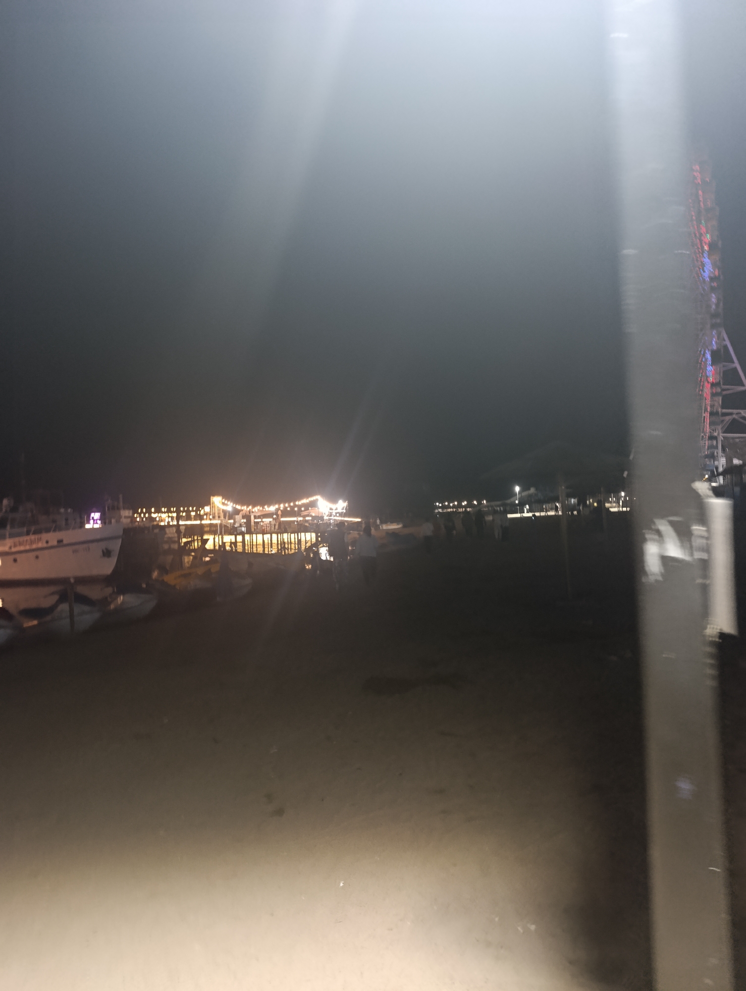 The image depicts a nighttime scene at a waterfront in Bosteri, Kyrgyzstan. A pier, illuminated by strings of lights, extends into the water. Several boats are moored near the shore, with one larger vessel on the left side of the frame. Silhouettes of people are visible on the pier and along the waterfront, suggesting a public area or gathering place. The overall atmosphere is dimly lit, with the lights from the pier and surrounding structures creating reflections on the water's surface. The sky is dark and overcast, with a faint glow emanating from the lights. In the far right of the image, a tall, narrow structure with colorful lights, possibly a Ferris wheel or tower, is partially visible. The ground in the foreground appears to be sandy or dirt.