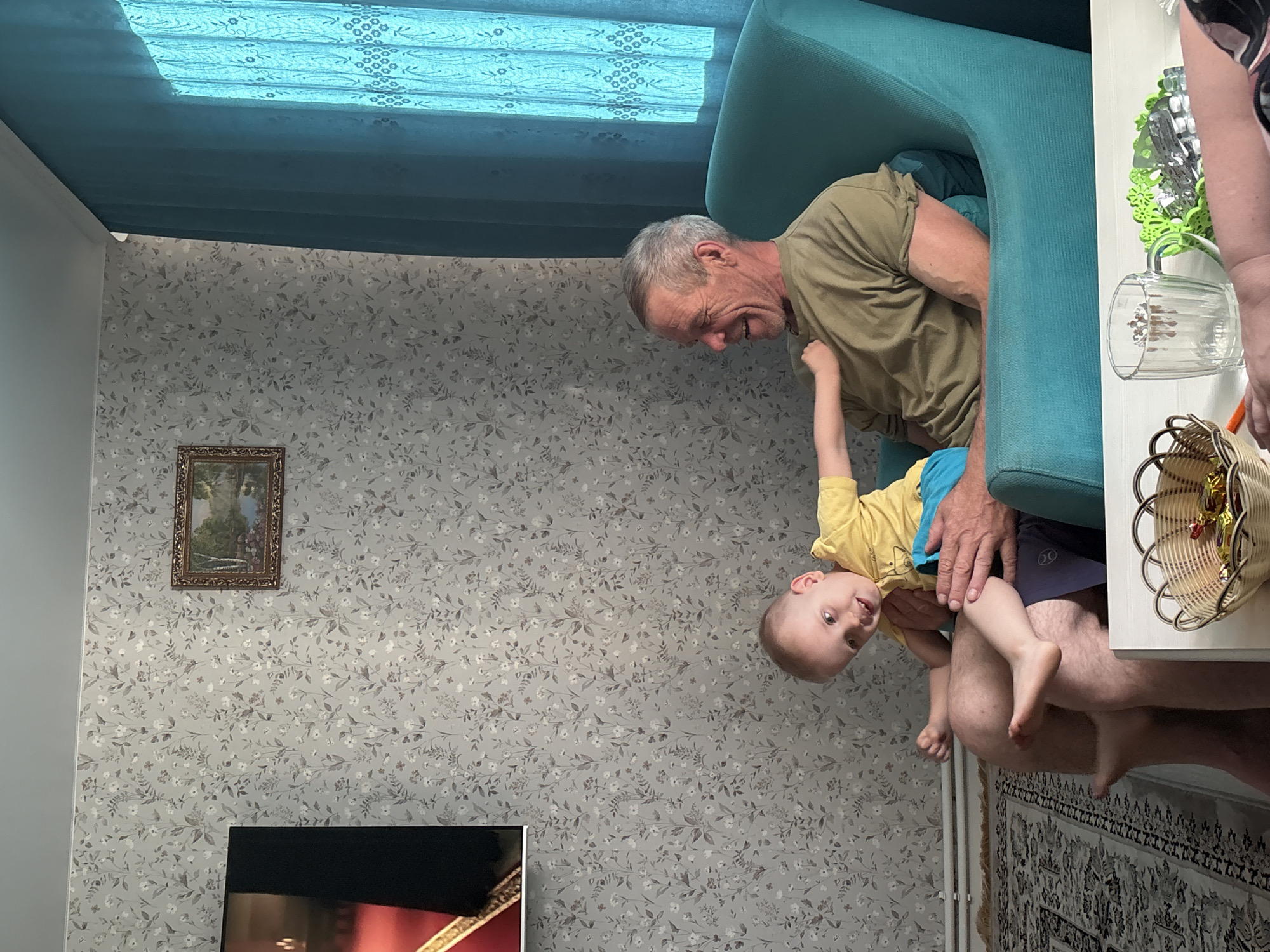Inside a room in what is likely a residential building, an older Caucasian male is seated in a turquoise armchair, smiling and looking down at a young child who is being held upside down in his lap. The child is wearing a yellow shirt and blue shorts. The room has floral wallpaper and a blue patterned window covering. A framed picture hangs on the wall. Part of a television is visible in the lower left corner. A small basket and a clear glass mug sit on a table next to the chair. The photo was taken in Chaikovsky, Russia.