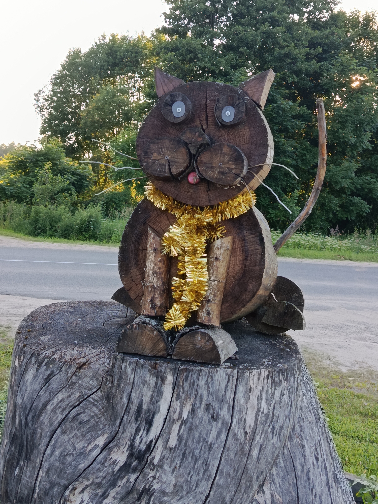 This is a medium shot, eye-level, outdoor photograph of a wooden cat sculpture situated on a tree stump. The sculpture is crafted from dark brown wood, resembling a rounded cat body with distinct ears, eyes, a nose, and a mouth. The eyes are made from circular metallic objects. A shiny gold tinsel garland is draped around the cat's neck. White wires protrude from the sides of its head, serving as whiskers, and a small red sphere is visible as its tongue. The sculpture's feet and tail are also made of wood. The cat is positioned on a large, weathered tree stump with visible wood grain and cracks. Behind the sculpture, a paved road runs horizontally, bordered by green grass and dense foliage. The background consists of trees, some with sunlight filtering through the leaves, suggesting it is late afternoon or early evening. The overall setting appears to be a rural roadside. This scene is located in the Мяжанскі сельскі Савет, Belarus.