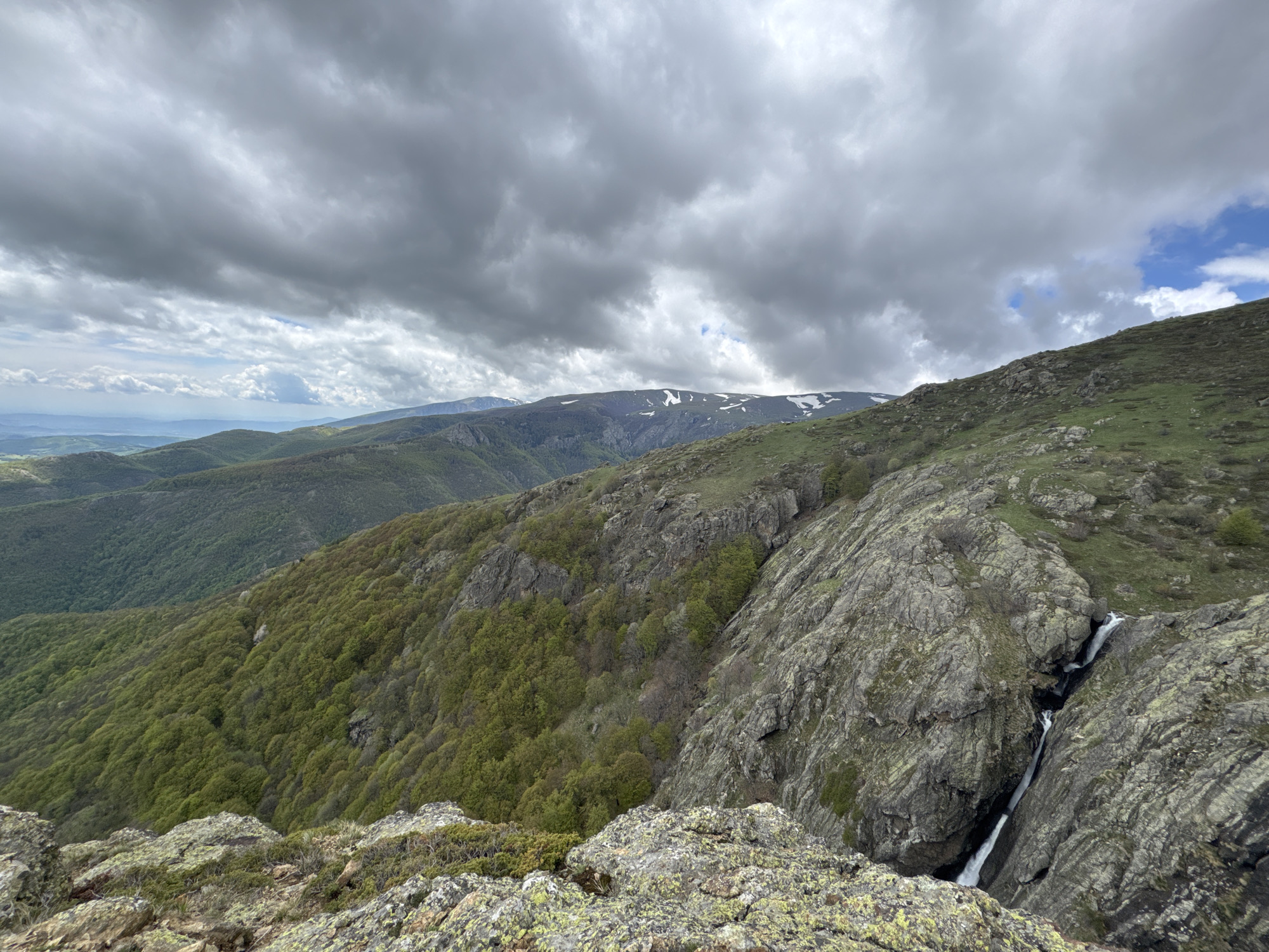 The image shows a mountainous landscape with a cloudy sky. In the foreground, there are lichen-covered rocks, leading to a steep, rocky cliff face with a small waterfall cascading down. To the left, a lush green hillside is visible, densely covered with trees. In the distance, other mountain ranges can be seen, with patches of snow on their peaks. The location is near Tazha, Bulgaria, based on the landscape characteristics of mountains and forests. There are no people or explicit actions visible in the scene.