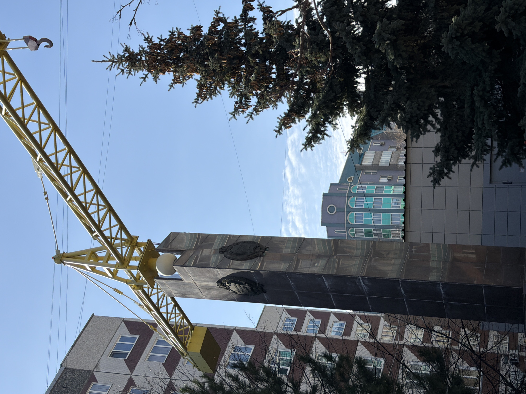 The image shows a monument in Krasnoyarsk, Russia, partially obscured by trees and viewed from a low angle. A yellow crane is also visible on the left side of the frame. The monument is a tall, dark structure with two circular emblems attached to its front. Buildings of varying architectural styles are visible in the background. The sky is blue with some light clouds. The photograph appears to have been taken during the day.