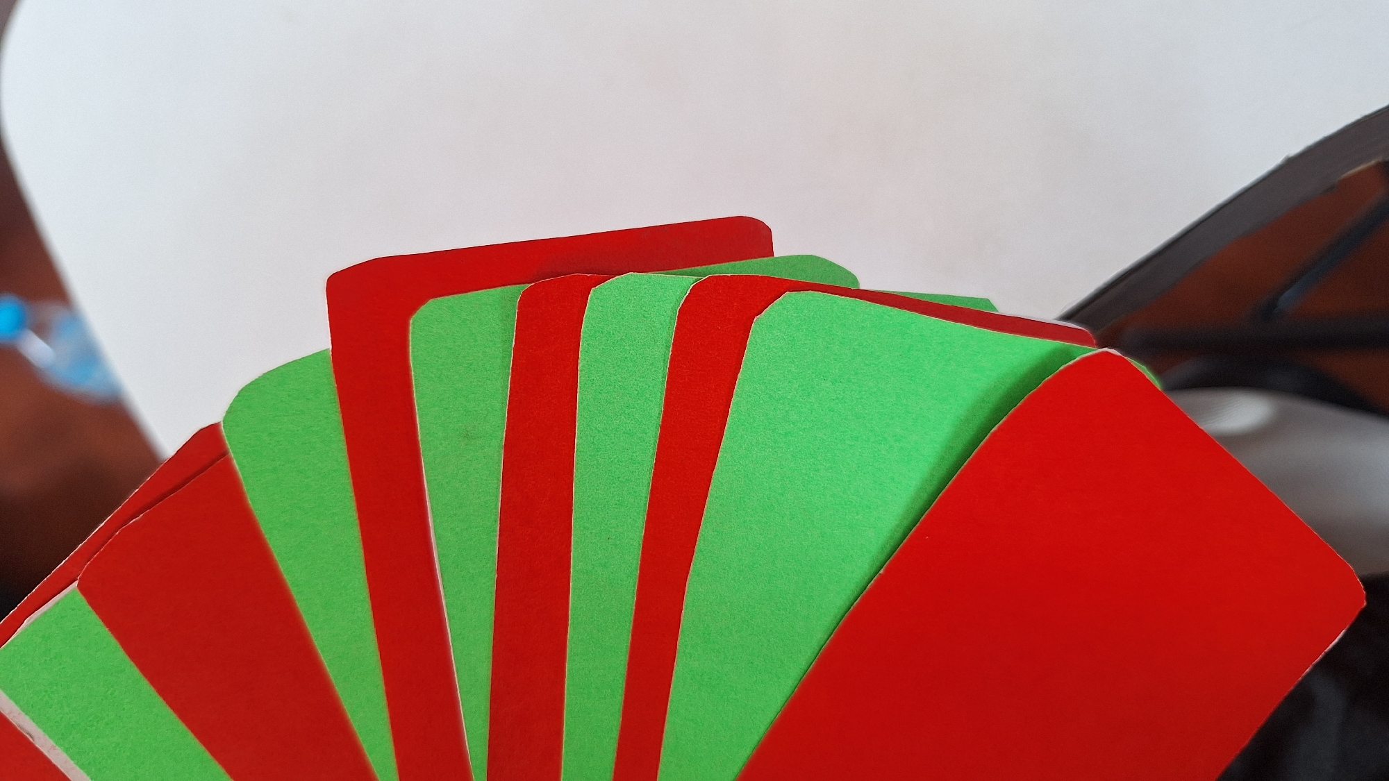 The image shows a fan of alternating red and green cards or paper. The cards are fanned out in a curved pattern, with the edges overlapping. The surface they are on appears to be a white table. In the background, a blurry blue object, possibly a bottle of water, is visible. The image is taken from a close-up, angled perspective, focusing on the cards. The scene is indoors and the lighting suggests it is daytime. There are no people or animals visible in the image. It is not possible to determine the location in Karawang, Indonesia based on the visual information provided.