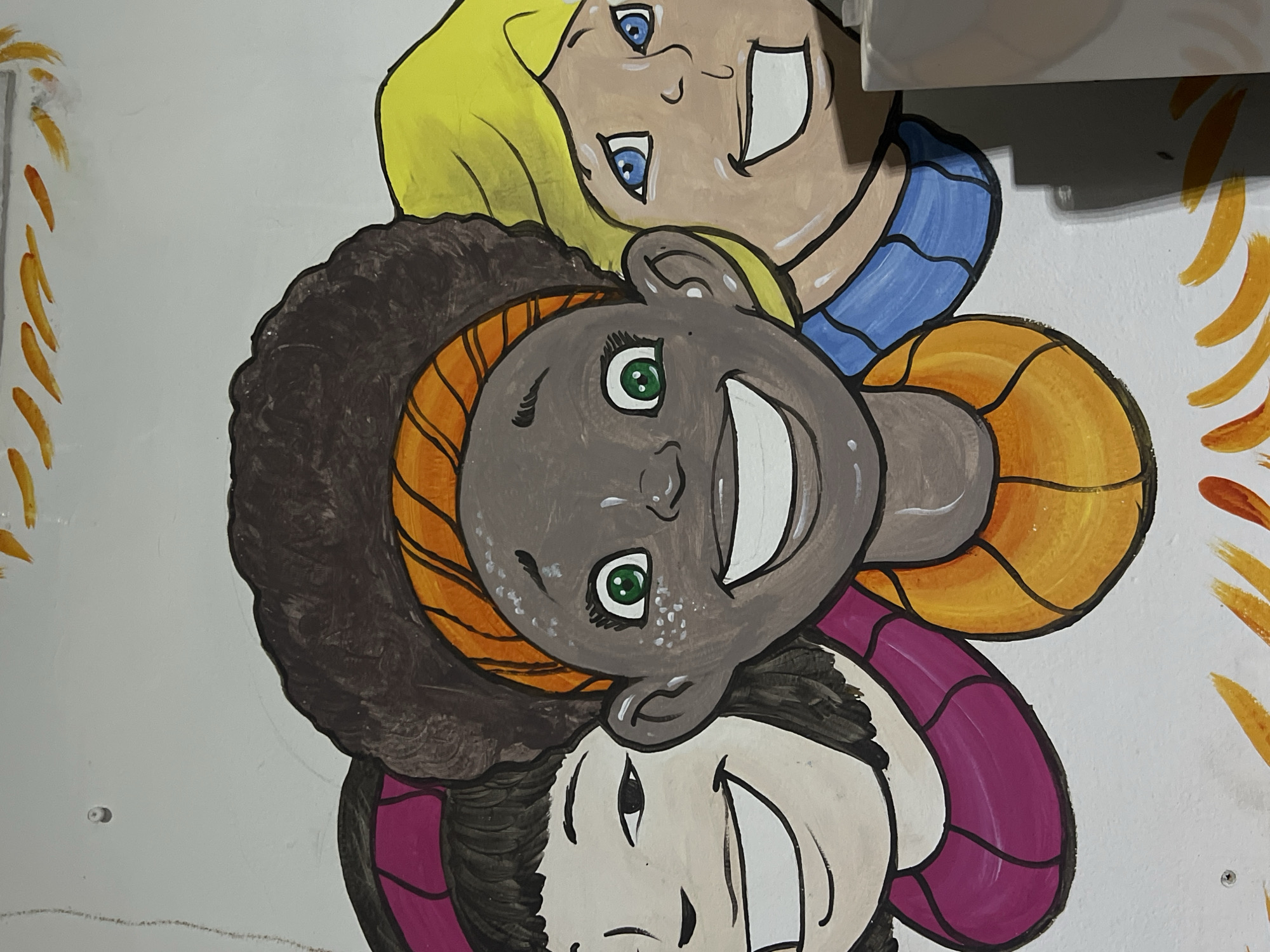A painting or mural features three smiling, cartoon-style faces stacked vertically on a white wall, presented horizontally due to the image's orientation. The characters appear to be children or young people with diverse features.
The uppermost face (when viewed as oriented in the photo) has light skin, blue eyes, and bright yellow hair. Below this, the middle face has dark skin, green eyes, and voluminous dark brown curly hair, adorned with an orange, striped hair accessory or scarf. The lowermost face has light skin, dark eyes, and dark hair, with a prominent purple, striped accessory or clothing element around its head or neck. All three faces are depicted with wide, open-mouthed smiles, showing white teeth, and have cheerful expressions.
The painting style uses bold black outlines and bright, distinct colors. Abstract, flame-like or feathery orange and yellow designs are painted along the edges of the wall, framing the faces. The background is a plain white wall. There are no activities depicted other than the subjects posing. Small details include minor scuffs on the wall and small screws or dots visible in the white space. No text is visible.