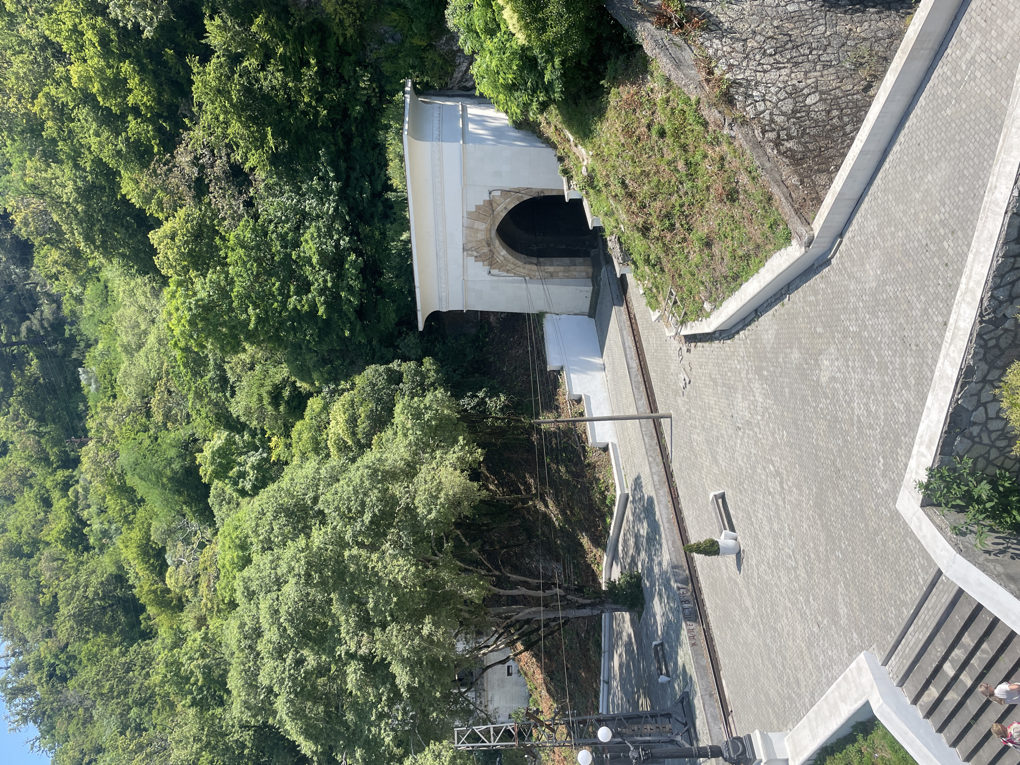 In Abaata, Abkhazia, a large, arch-shaped tunnel entrance, framed by light-colored stone and topped with a white facade, dominates the right side of the image. The tunnel is dark inside. The entrance is situated on a grassy, sloped embankment covered in vegetation. To the left of the tunnel, a paved area transitions to a railway track. A lone tree with a thick trunk and expansive branches stands in the middle ground, casting shadows. Further left, a black lamppost with two lights and a metal structure are visible. The ground is paved with gray interlocking bricks. In the foreground, a concrete staircase leads down, with two individuals partially visible at the bottom. The scene is set against a backdrop of a dense, verdant forest covering a steep hillside under a clear blue sky.