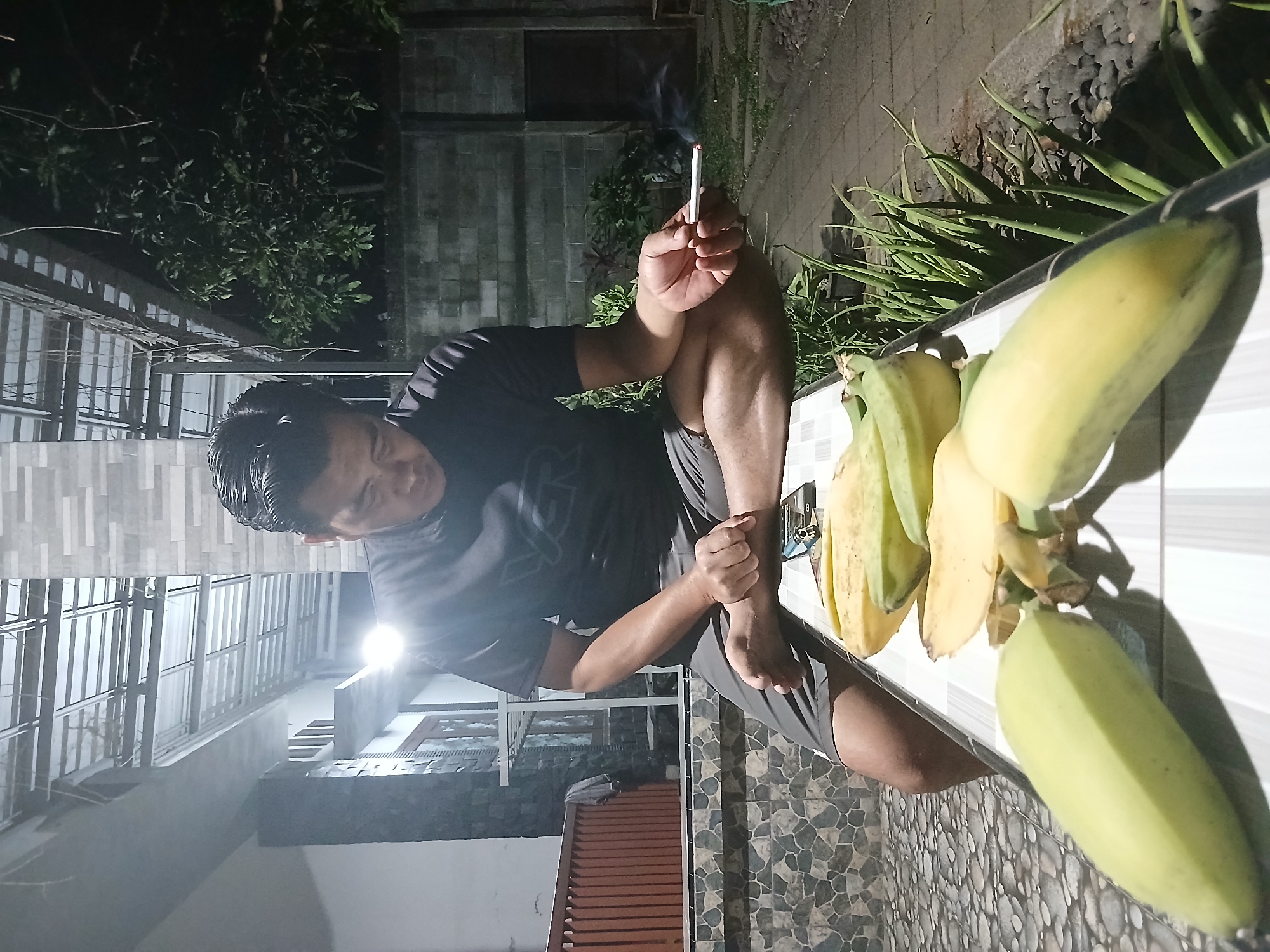 A man sits cross-legged on a tiled ledge at night in Sekarputih, Indonesia. He is wearing a dark t-shirt and shorts, and holding a cigarette that is lit and emitting smoke in his right hand. In front of him on the ledge, there are several bananas, some of which appear to be partially peeled. The man is looking down at his cigarette and the bananas. The background shows a building with a corrugated roof, a stone wall, and some greenery. The scene appears to be outdoors, possibly in a backyard or patio area.
