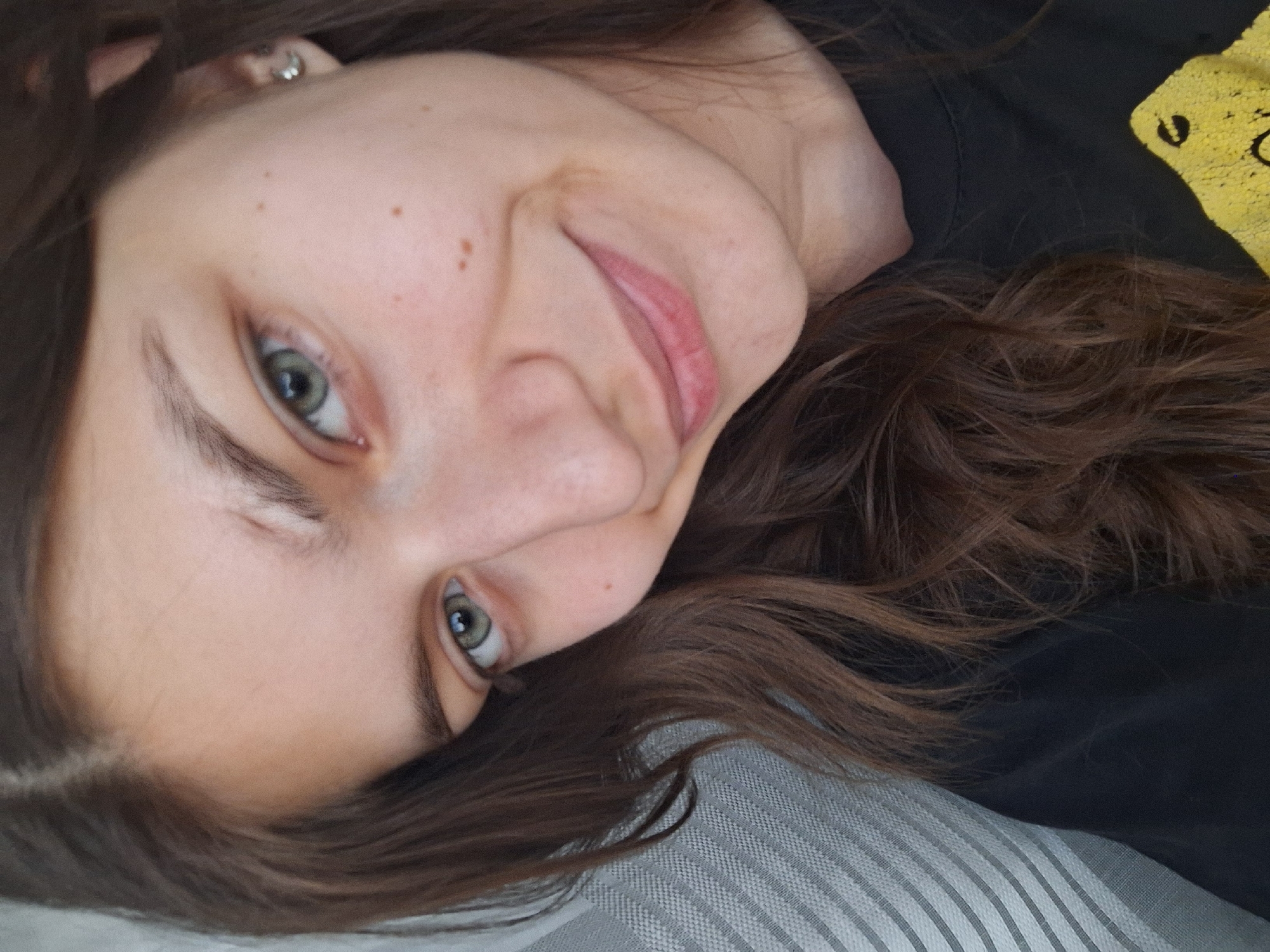 A close-up, tilted portrait of a light-skinned person with brown hair and green eyes. The person is lying on a striped surface, possibly a pillow or blanket. They are wearing a black shirt with yellow designs on the upper right. The person is smiling subtly. The background is blurred. There is no indication of this image being taken in Panagyurishte, Bulgaria, nor any identifiable geographical location.